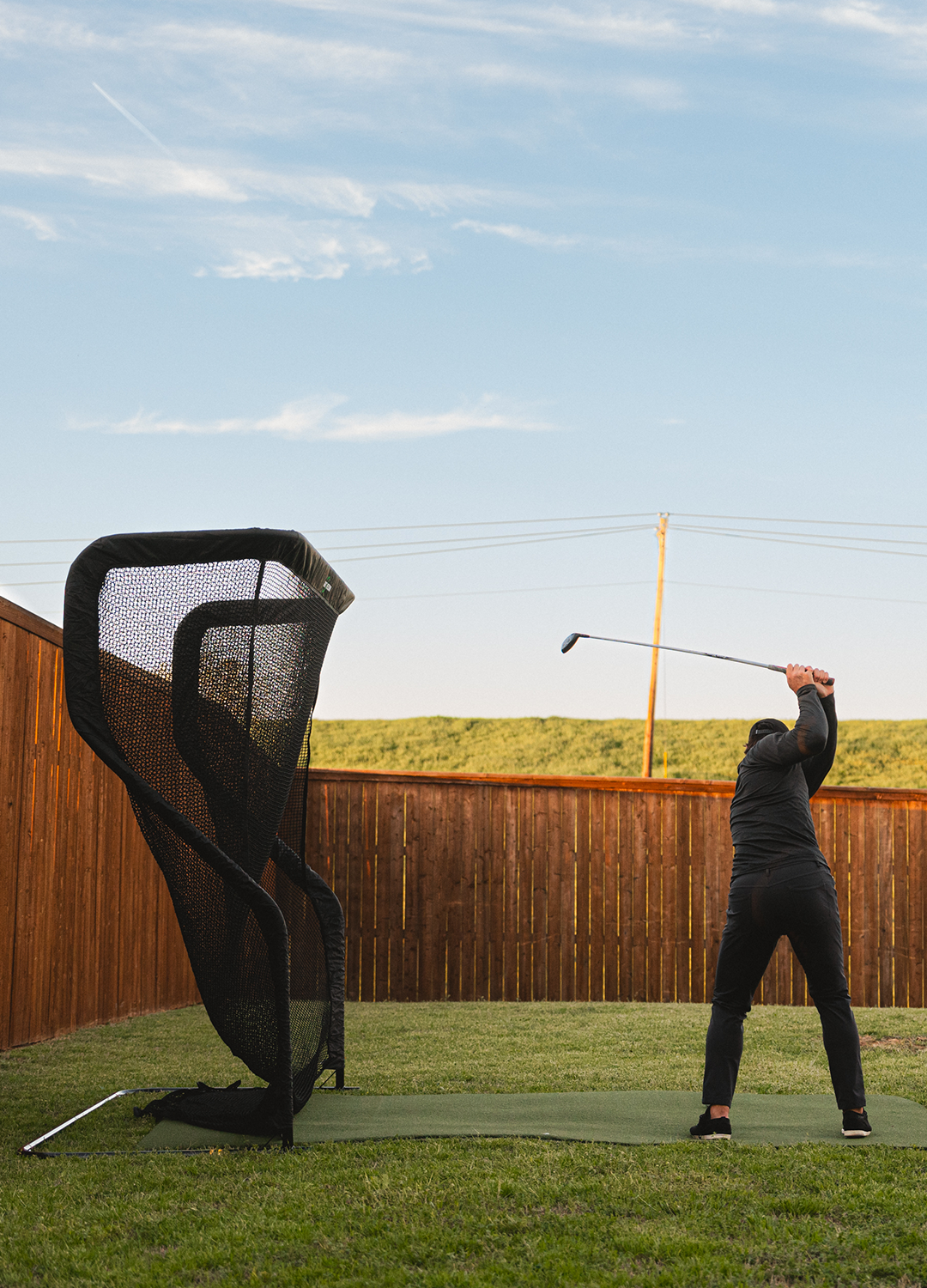 A person swings a golf club in a backyard practice area with a net.