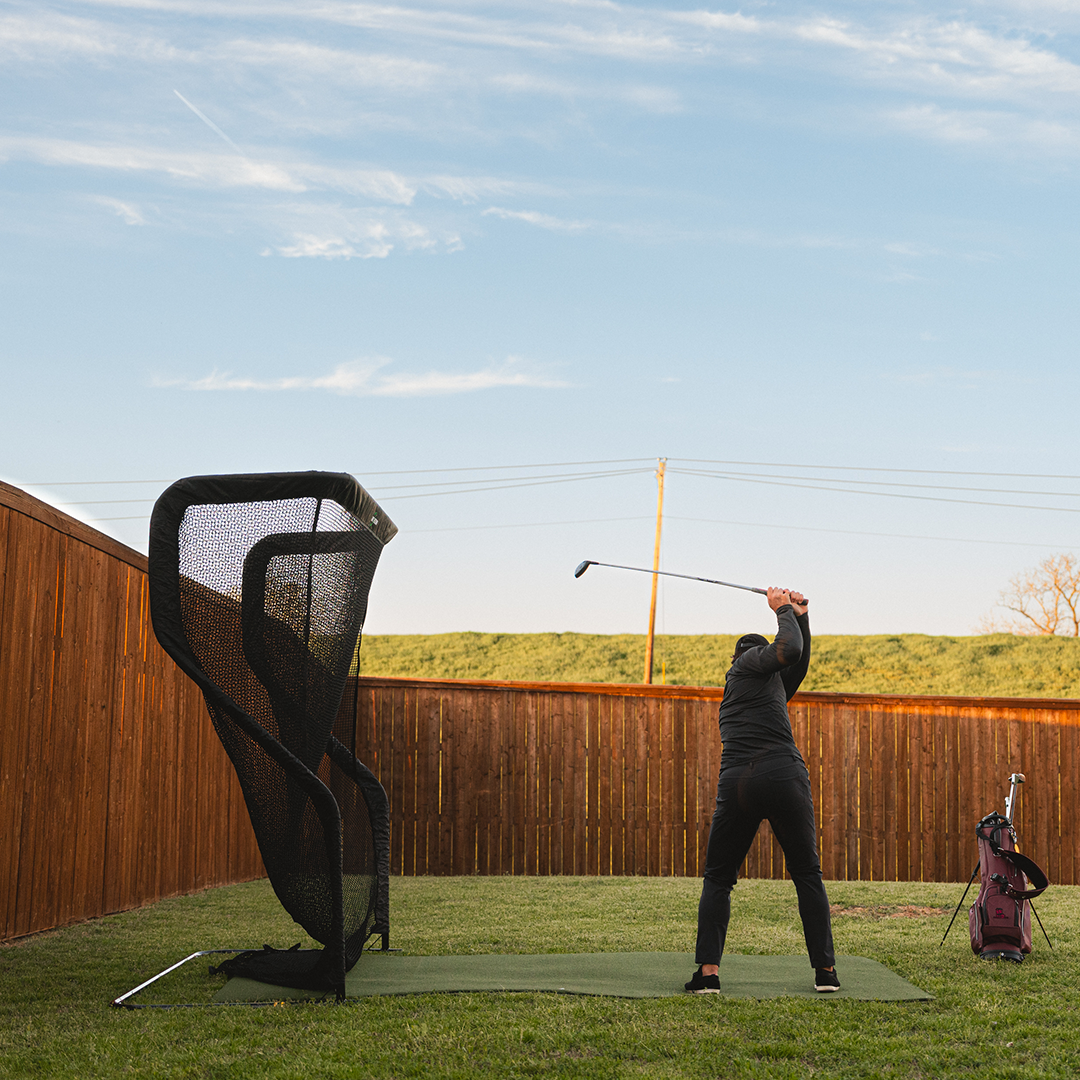 Person practicing golf swings in a backyard using a practice net, with a golf bag nearby. Fence and open sky in the background.