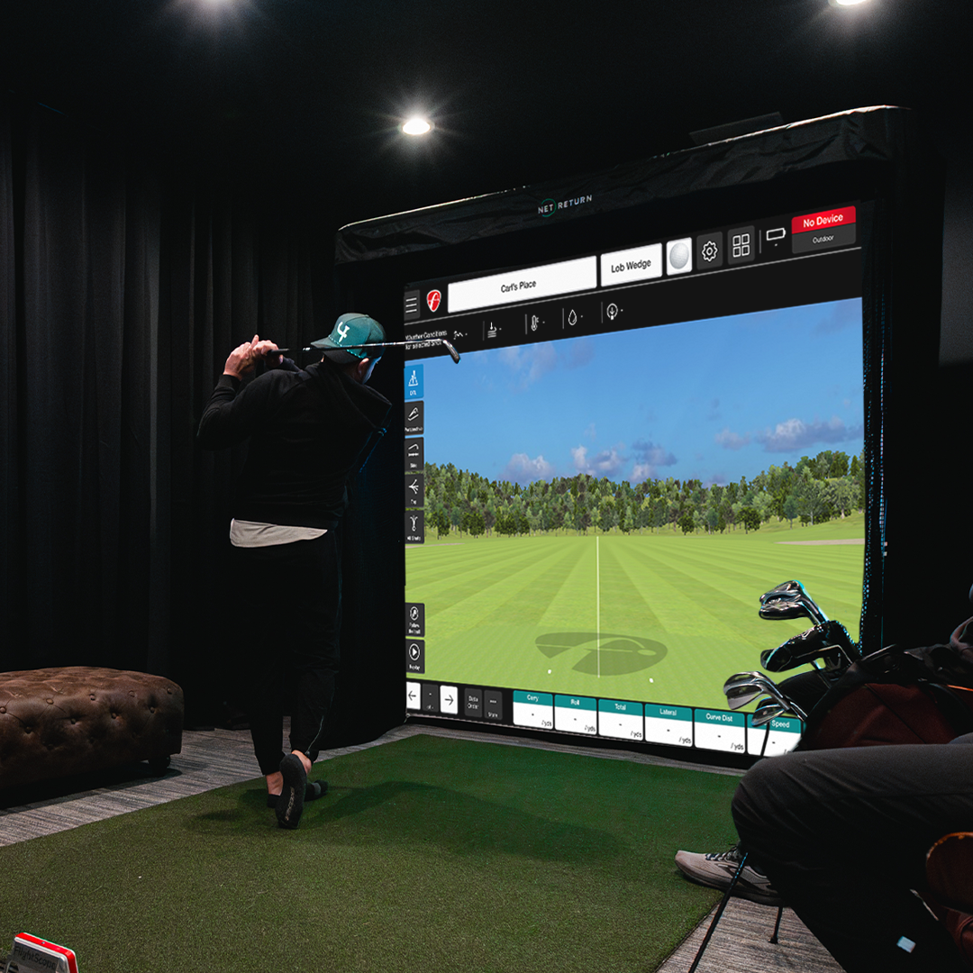 Person playing golf on an indoor virtual simulator, swinging a club in front of a large screen displaying a golf course. Golf bag and clubs are visible on the right.