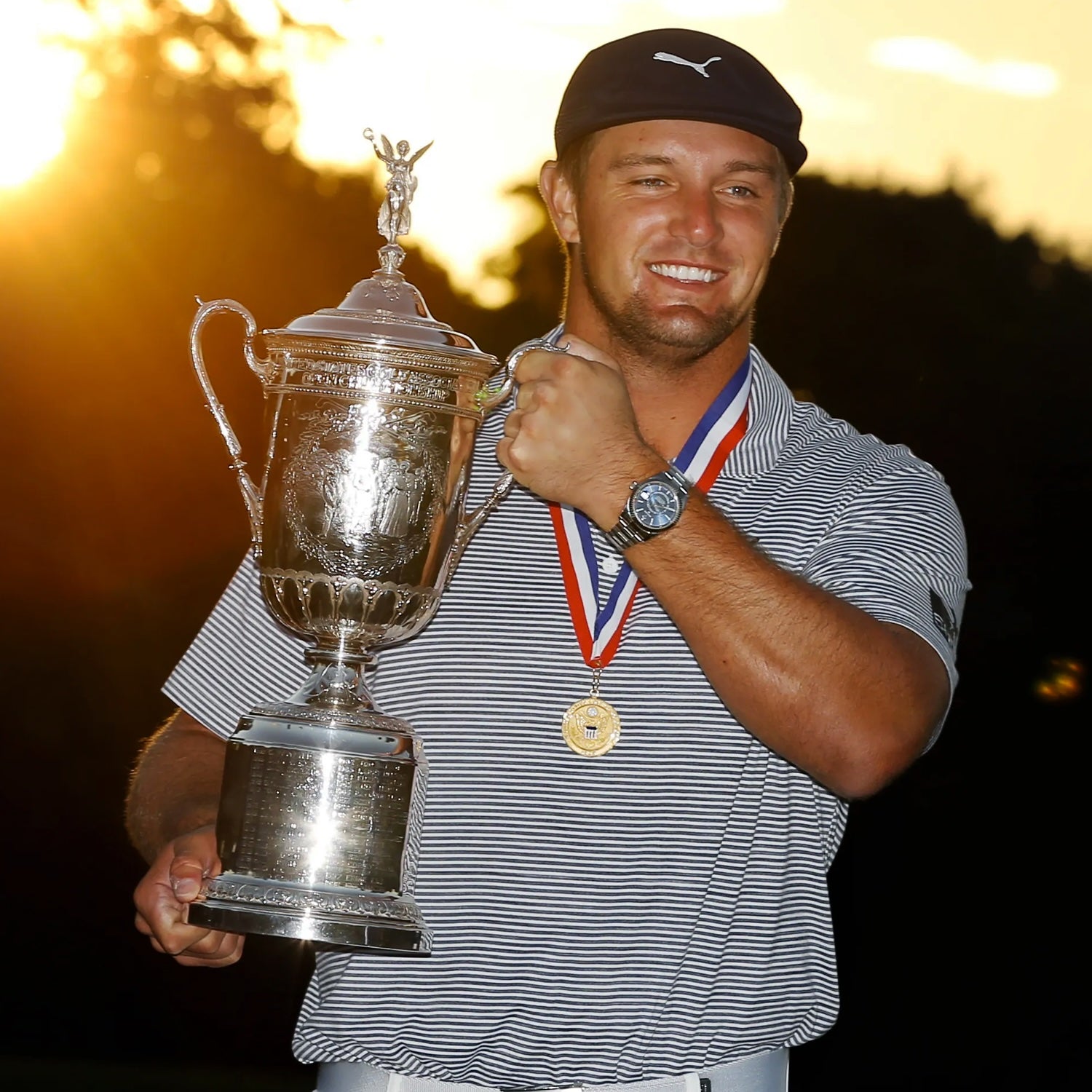 A golfer smiles and holds a trophy, wearing a medal and cap, with the sun setting in the background.