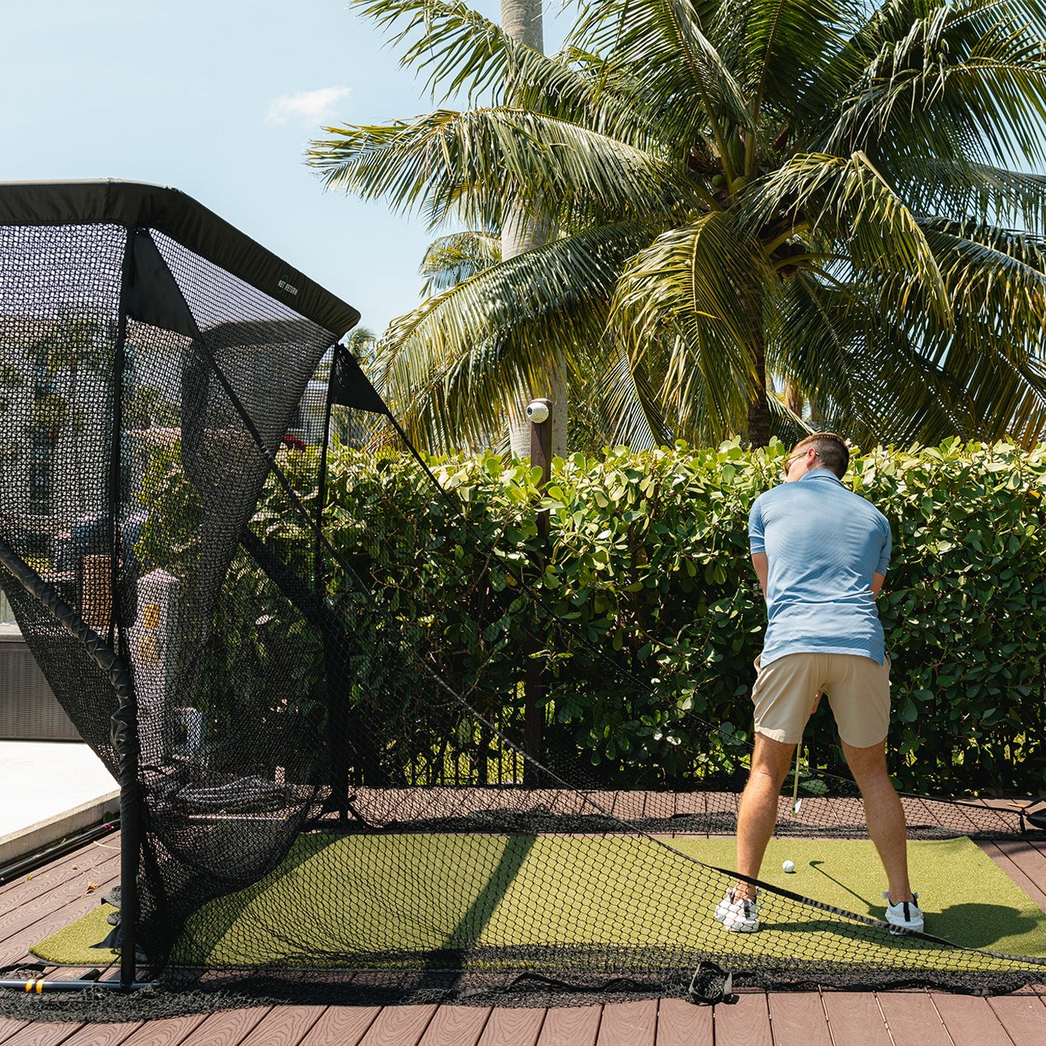 A man in a blue shirt and shorts practices golf swings on artificial grass with a net enclosure, surrounded by palm trees and greenery.