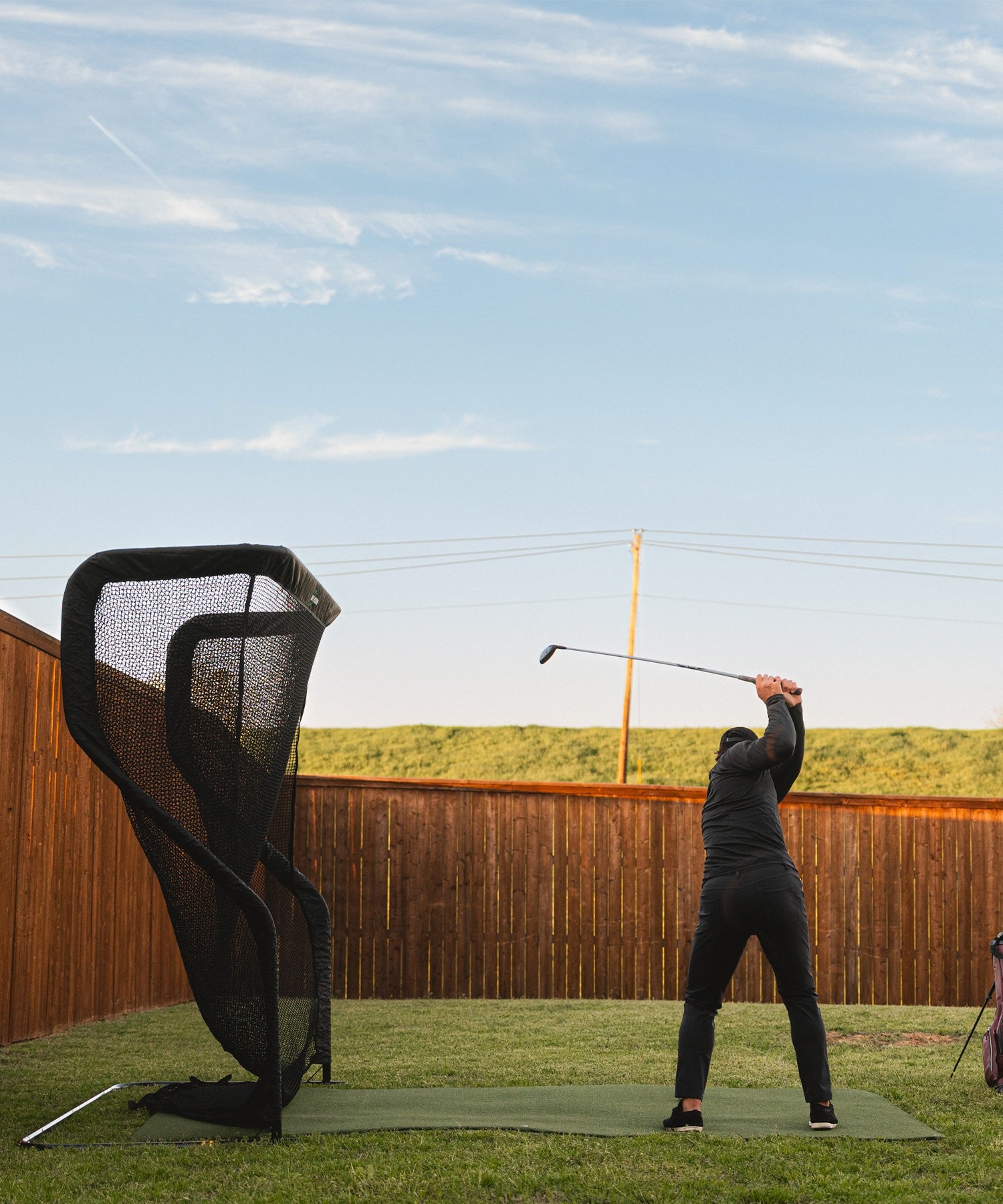 Person swinging a golf club in a backyard with a practice net under a clear sky.