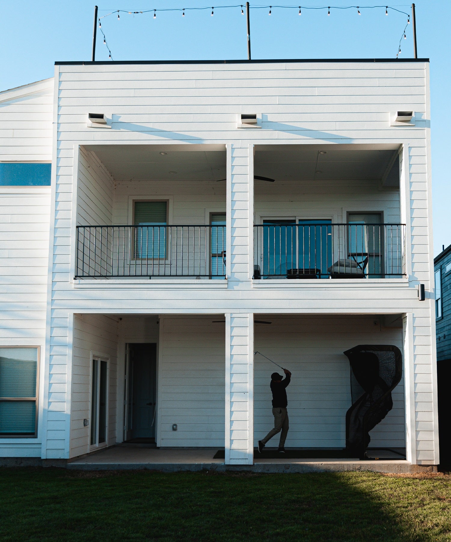 Person practicing golf swing on a patio of a two-story white house with balconies and string lights.
