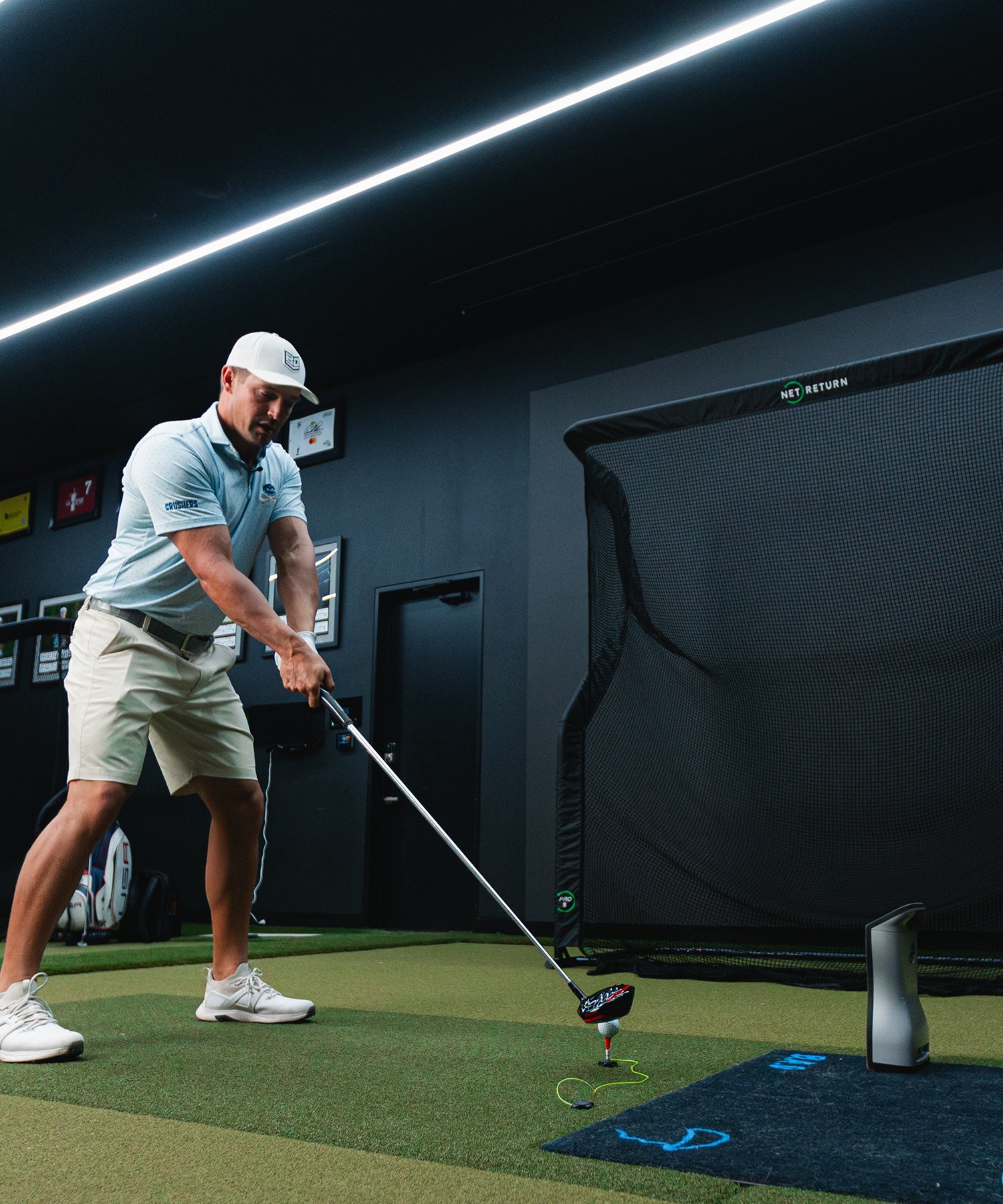 A golfer prepares to swing in an indoor practice area with a net and a monitor.