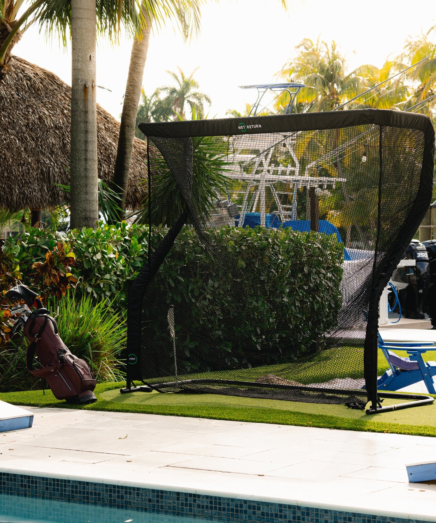 A golf practice net set up by a poolside, with a golf bag nearby. Tropical plants and a boat are in the background.