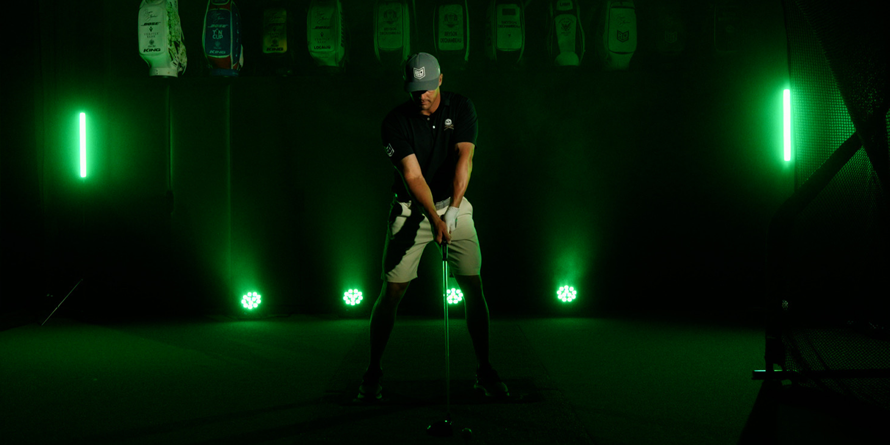 A golfer in a dark room prepares to swing, surrounded by green lights and various hanging golf bags.