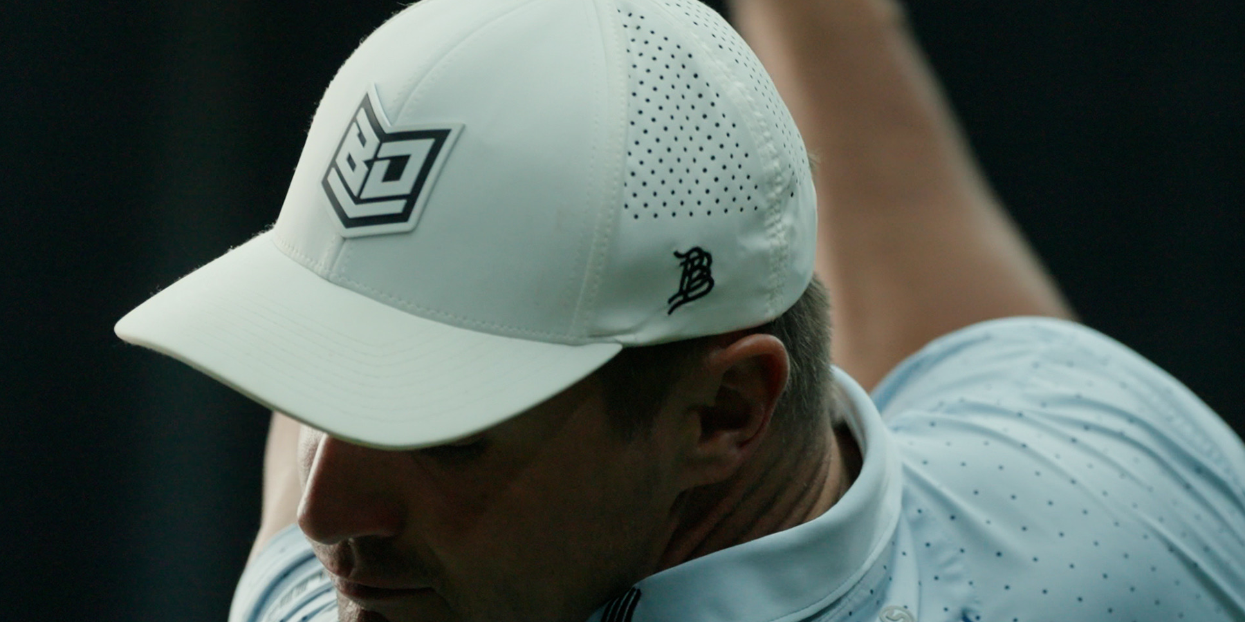 A person wearing a white baseball cap with a logo and a light blue shirt is seen from the side. The focus is on the cap.