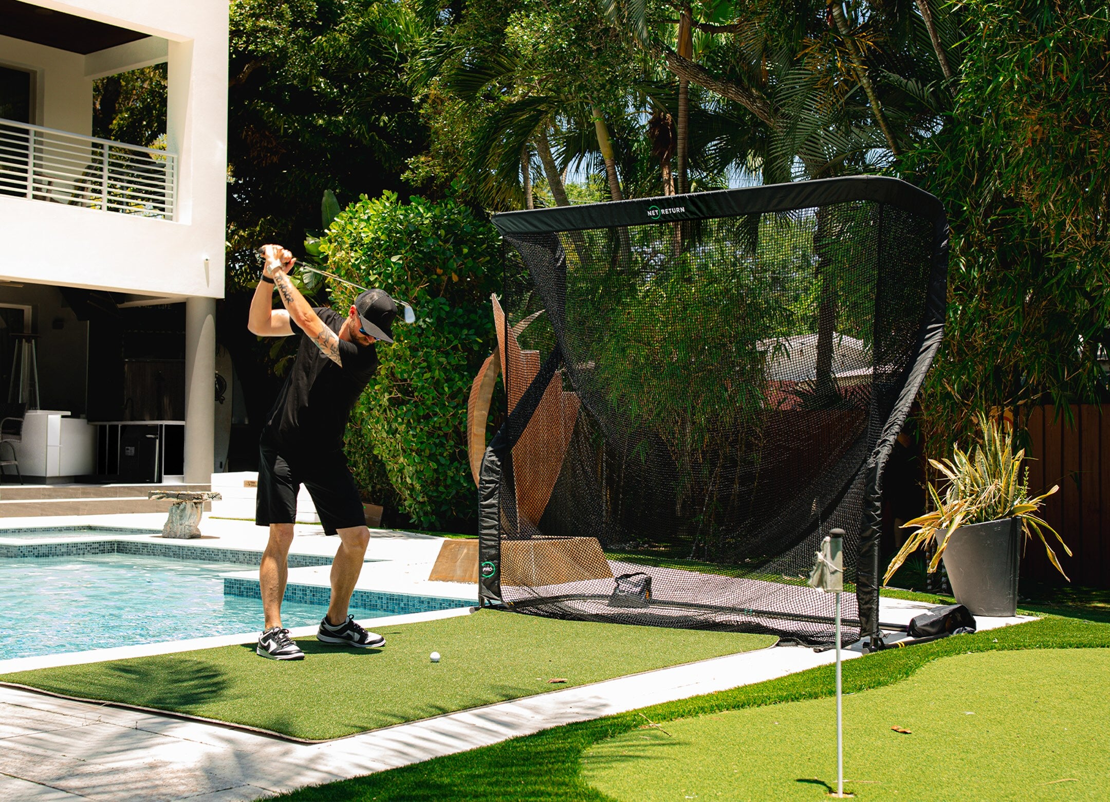 A person practices golf swings in a backyard with a net set up near a pool.