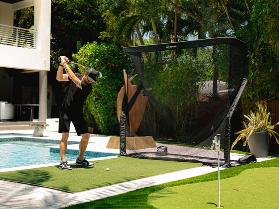 Person practicing golf swing in backyard using a golf net near a pool, surrounded by greenery.