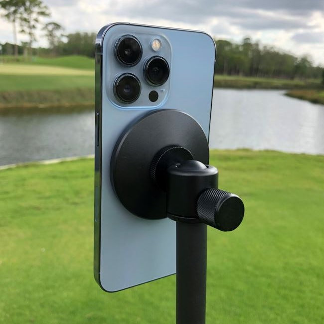 A smartphone is mounted on the GPOD by GPOD GOLF, set up outdoors beside a pond and grassy area with trees in the background.