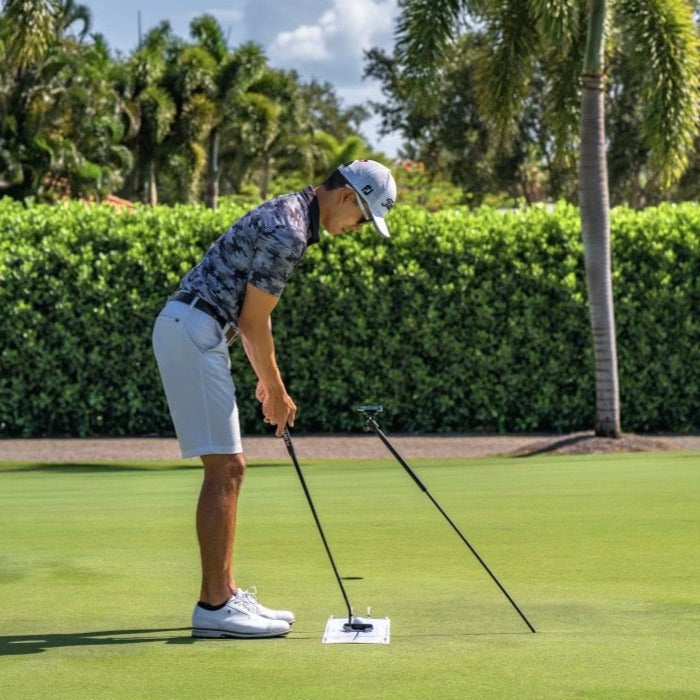 A golfer practices putting on the green using two alignment rods and the GPOD by GPOD GOLF MagSafe Training Aid, surrounded by palm trees and lush greenery.