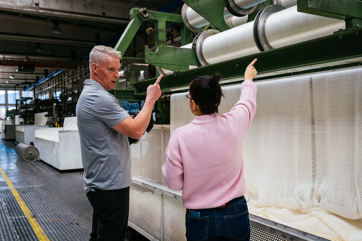 Two people discussing and pointing at machinery in a textile factory. One is holding a tablet.