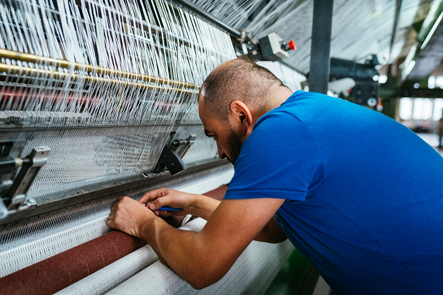 Man in blue shirt working at a loom, adjusting threads on a weaving machine in a textile factory.