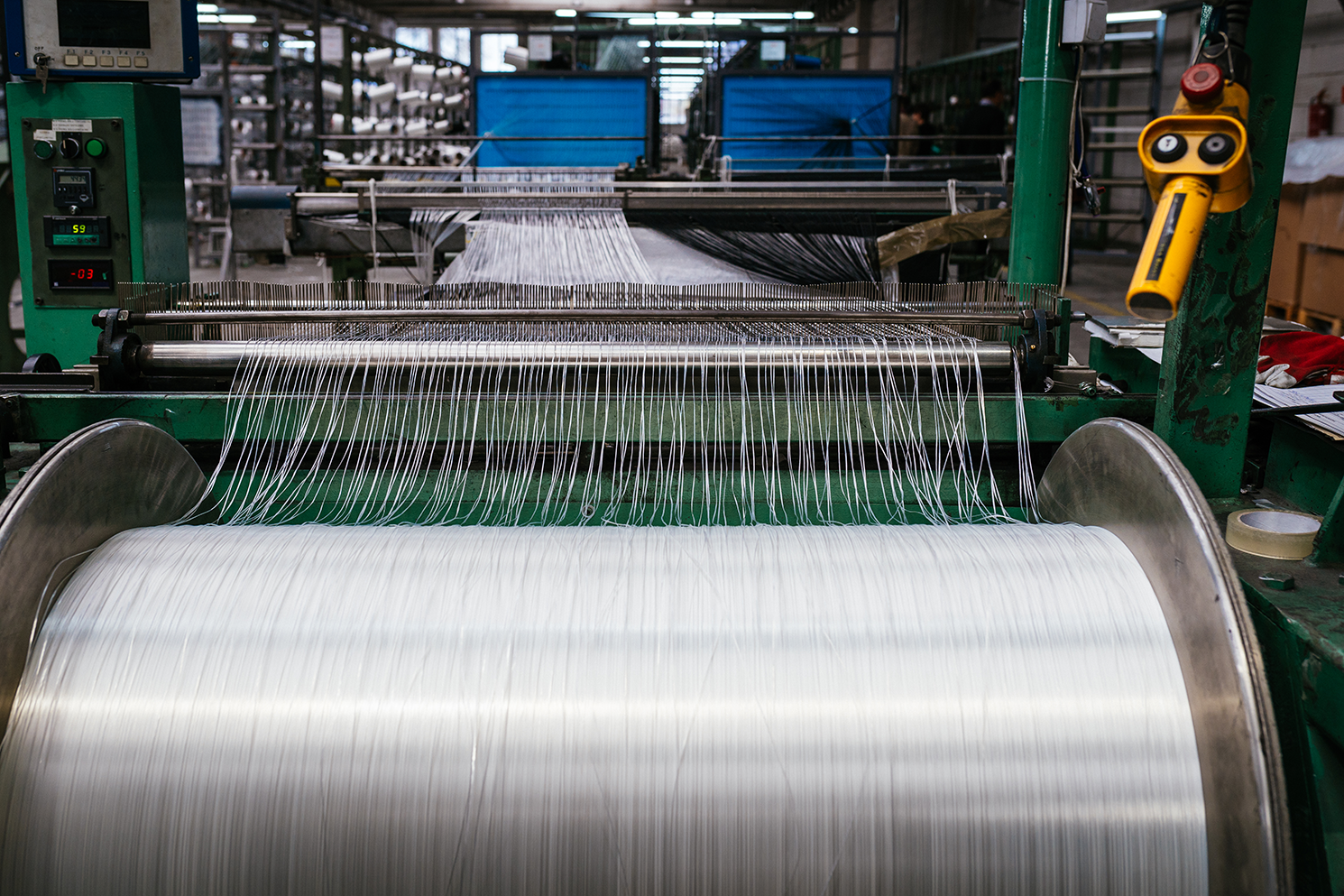 Industrial textile weaving machine with threads on rolls and fabric being woven in a factory setting.