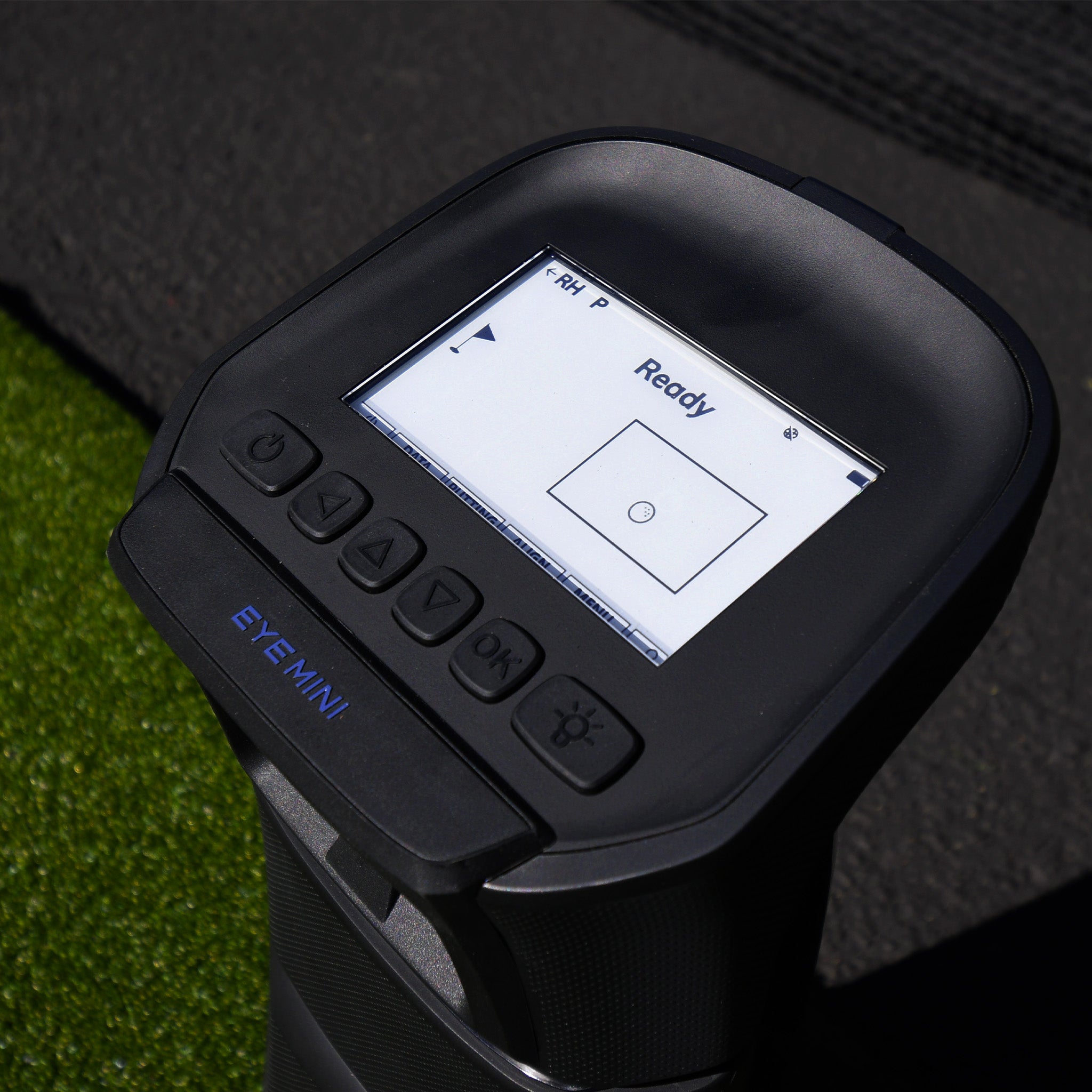Close-up of a black device labeled "EYE MINI" with a display screen showing "Ready" and a target. Five buttons are below the screen. Grass is visible in the background.
