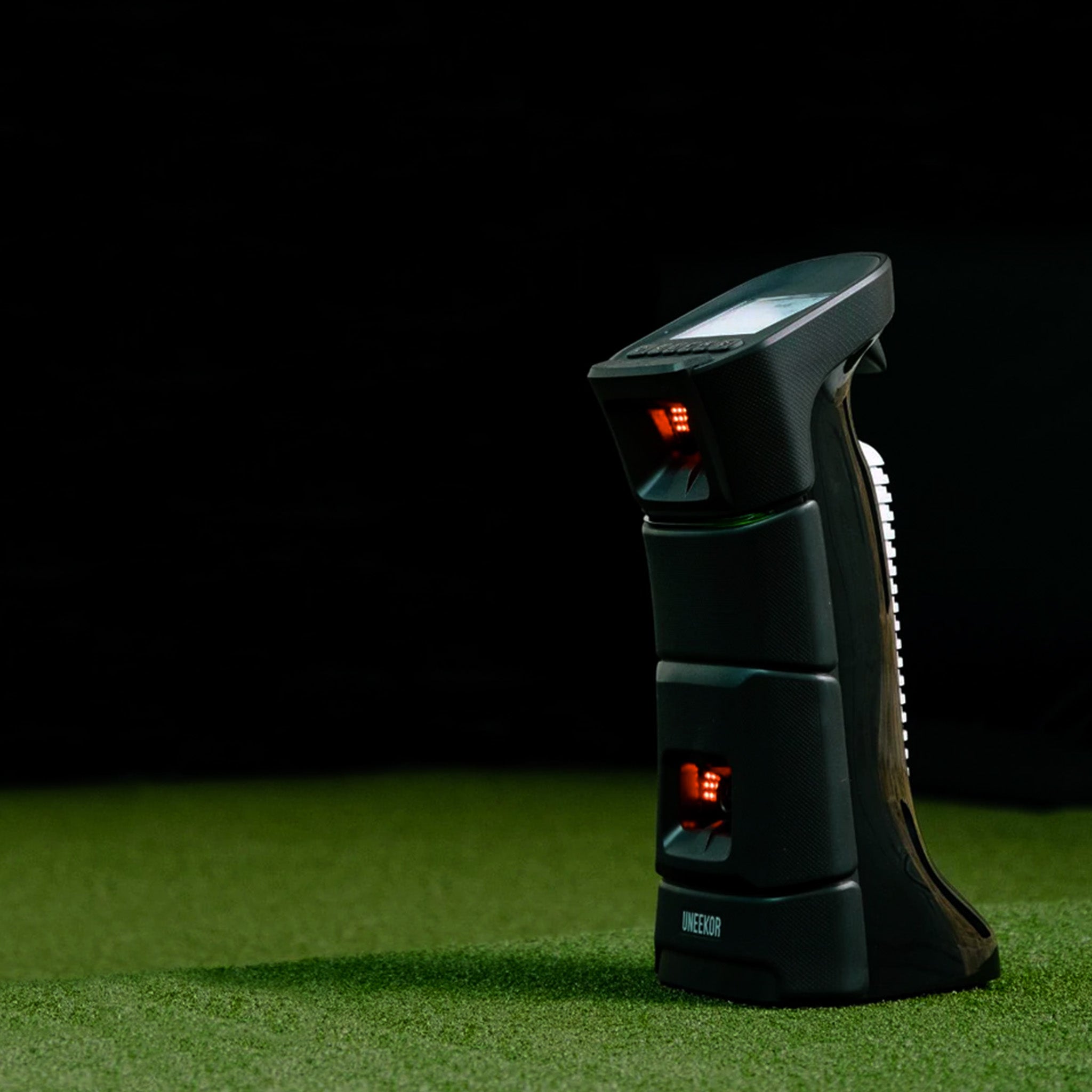 A black, upright electronic device with a digital screen stands on grass against a dark background.