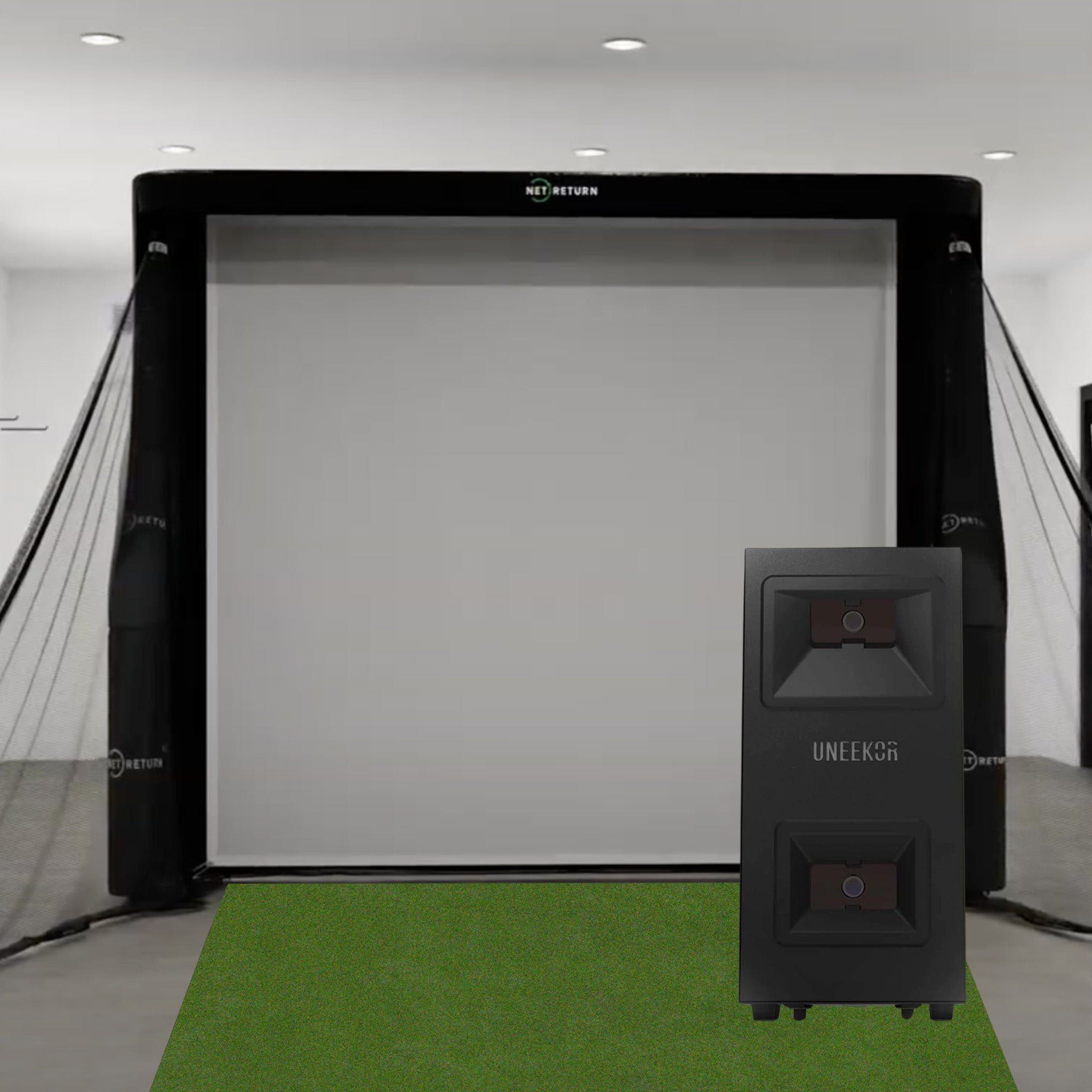 Indoor golf simulator setup with a large screen, netting on the sides, and a UNEEKOR device in front on a green mat.