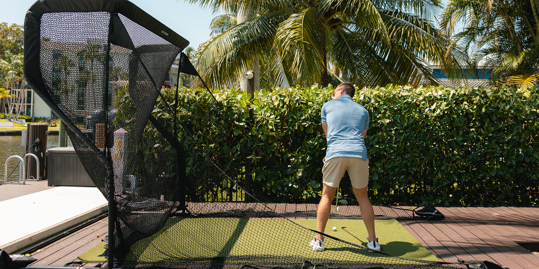 A person practices golf swings in a backyard setup with a net and artificial turf, surrounded by greenery.