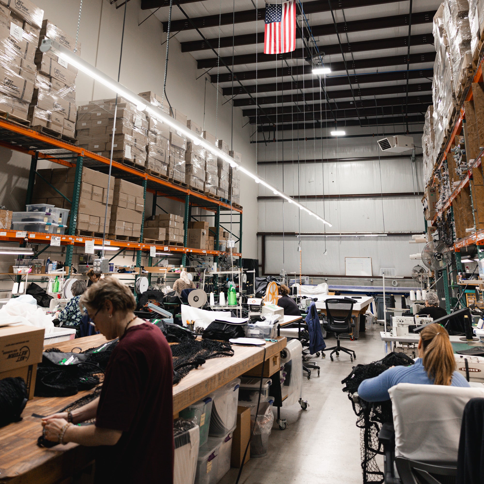 A warehouse interior with stacks of boxed items on shelves. People are seated at tables working with fabric materials under bright overhead lighting. An American flag hangs from the ceiling.