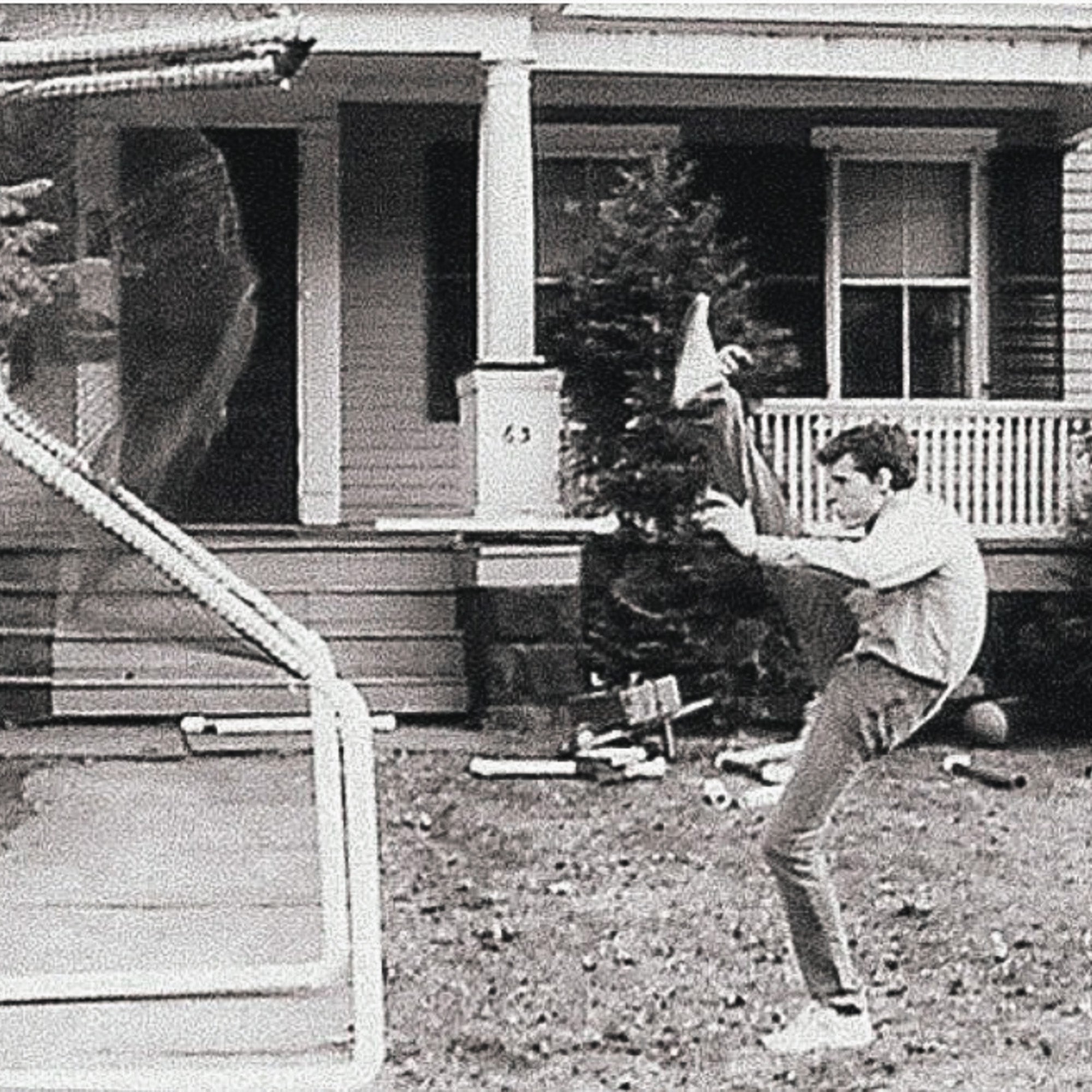 A man kicks high in front of a house, appearing to strike a falling basketball hoop. Debris is scattered on the lawn.