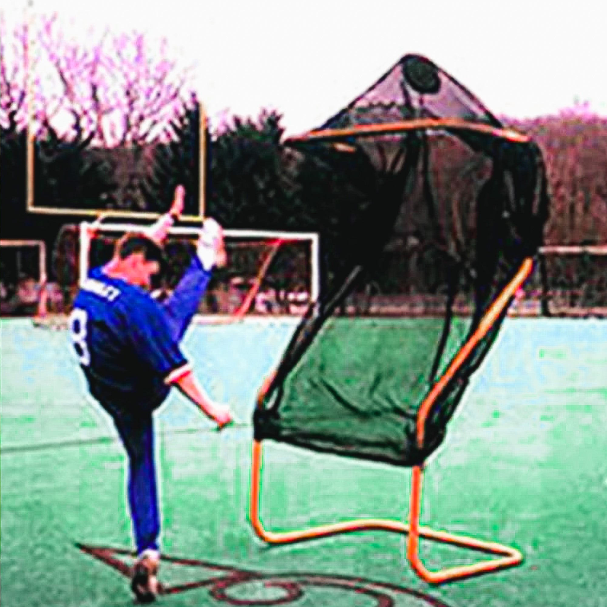 Person wearing a blue jersey practices kicking a football towards a large net on a green field. Multiple goalposts are in the background.