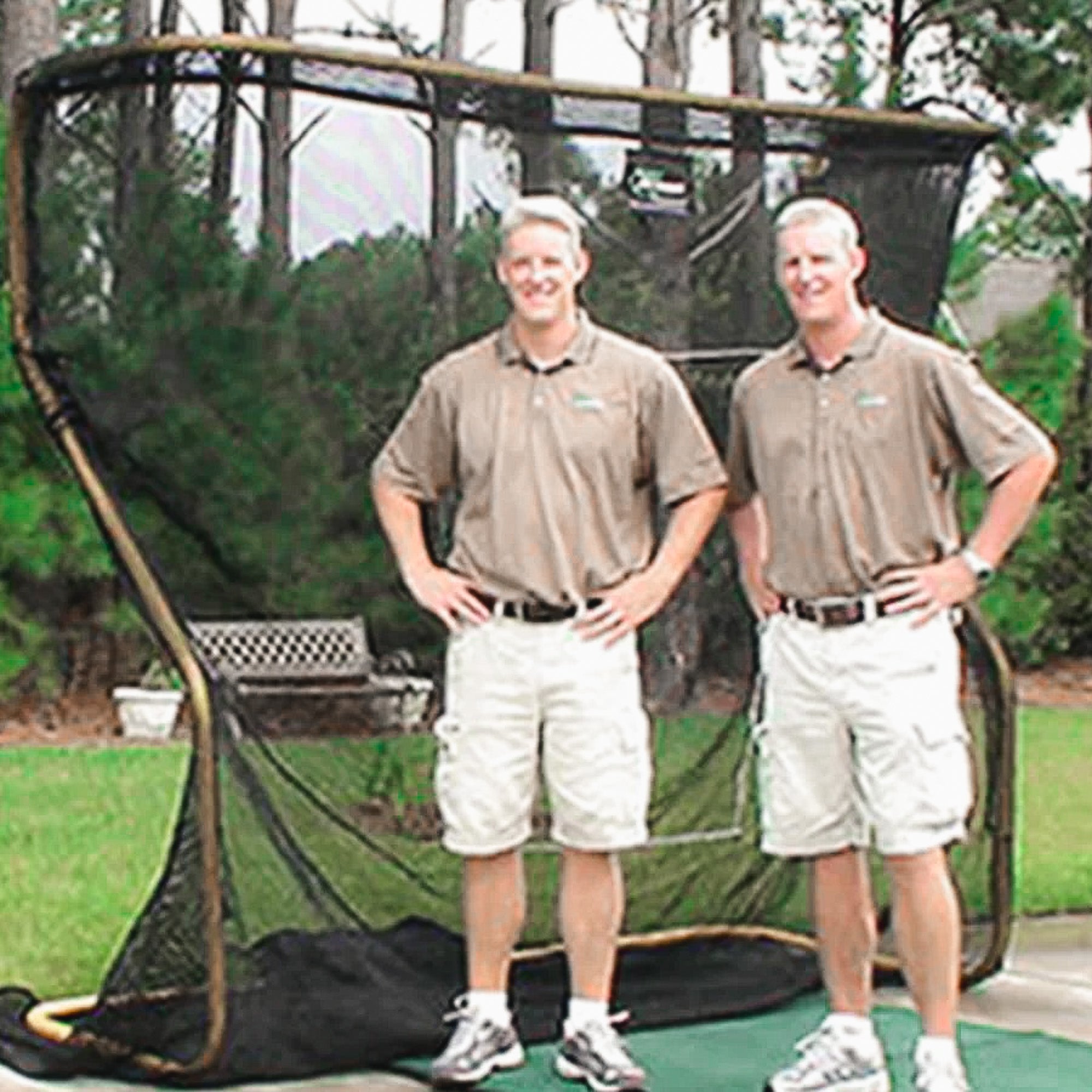 Two people in matching beige shirts and shorts stand in front of a large black net structure outdoors. Trees are visible in the background.