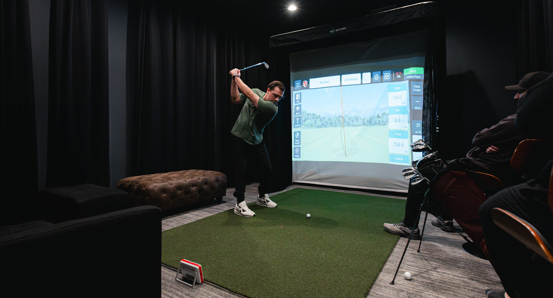 Person practicing golf swings indoors using a golf simulator screen, with others seated nearby observing.
