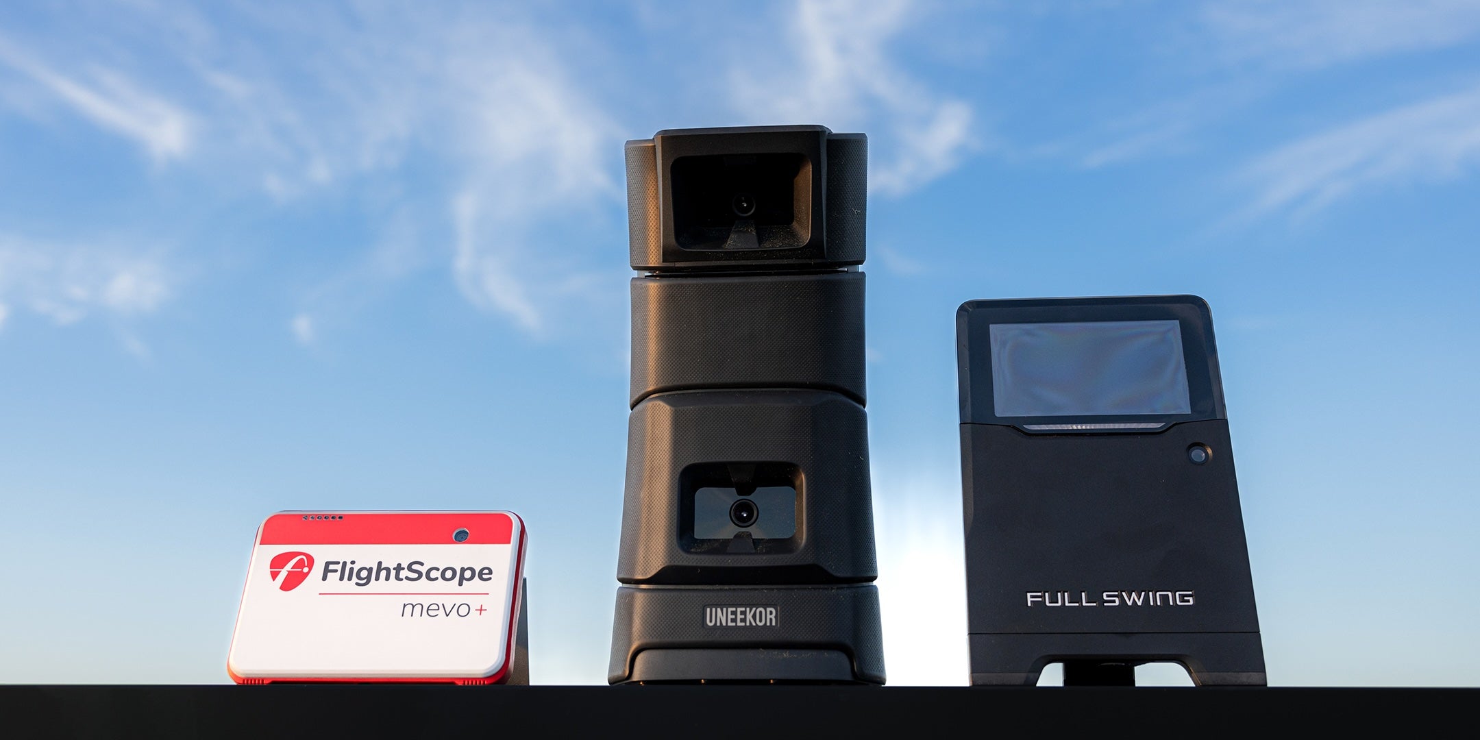 Three golf launch monitors—FlightScope Mevo+, Uneekor, and Full Swing—are displayed against a blue sky.