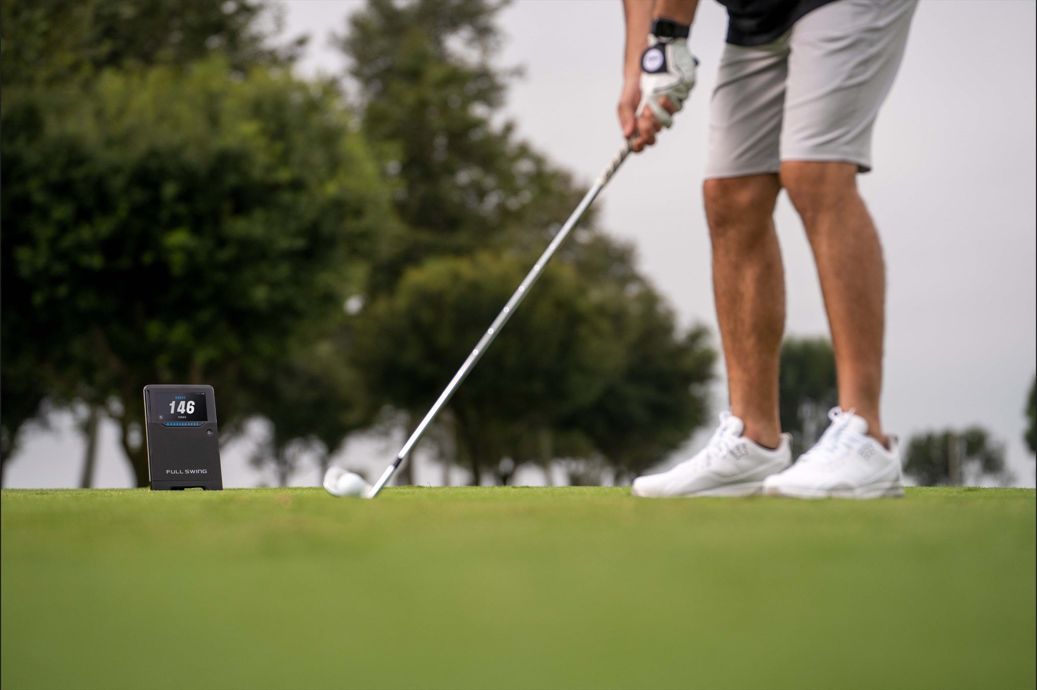 A golfer in shorts and sneakers prepares to putt on a green, with a digital device displaying "146" placed nearby. Trees are visible in the background.