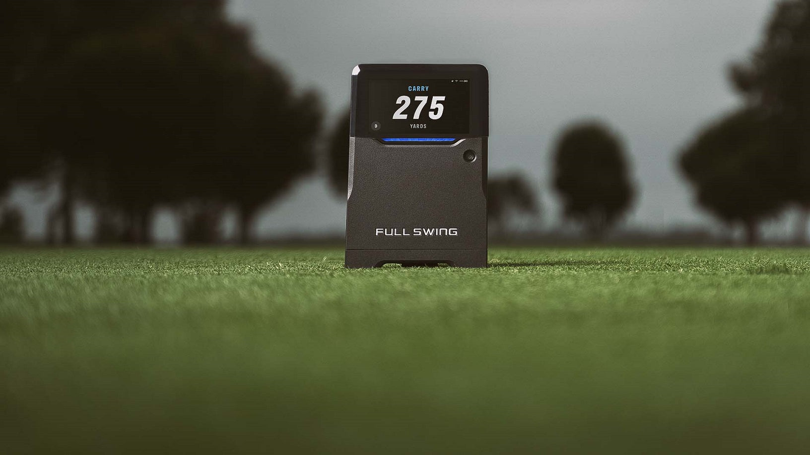 A golf launch monitor on a grassy field displays the number 275. Trees are visible in the blurred background.