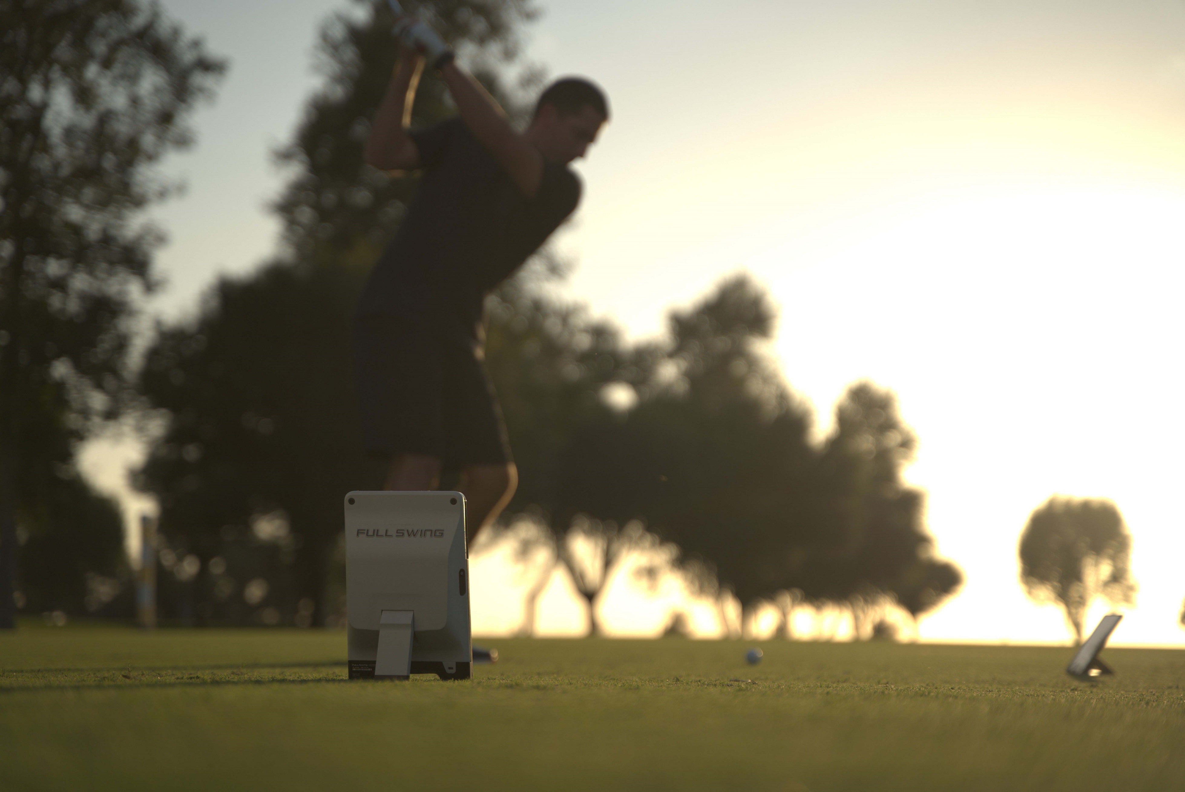 A golfer swings on a sunny golf course with trees in the background. A golf simulator device labeled "Full Swing" is positioned on the grass.