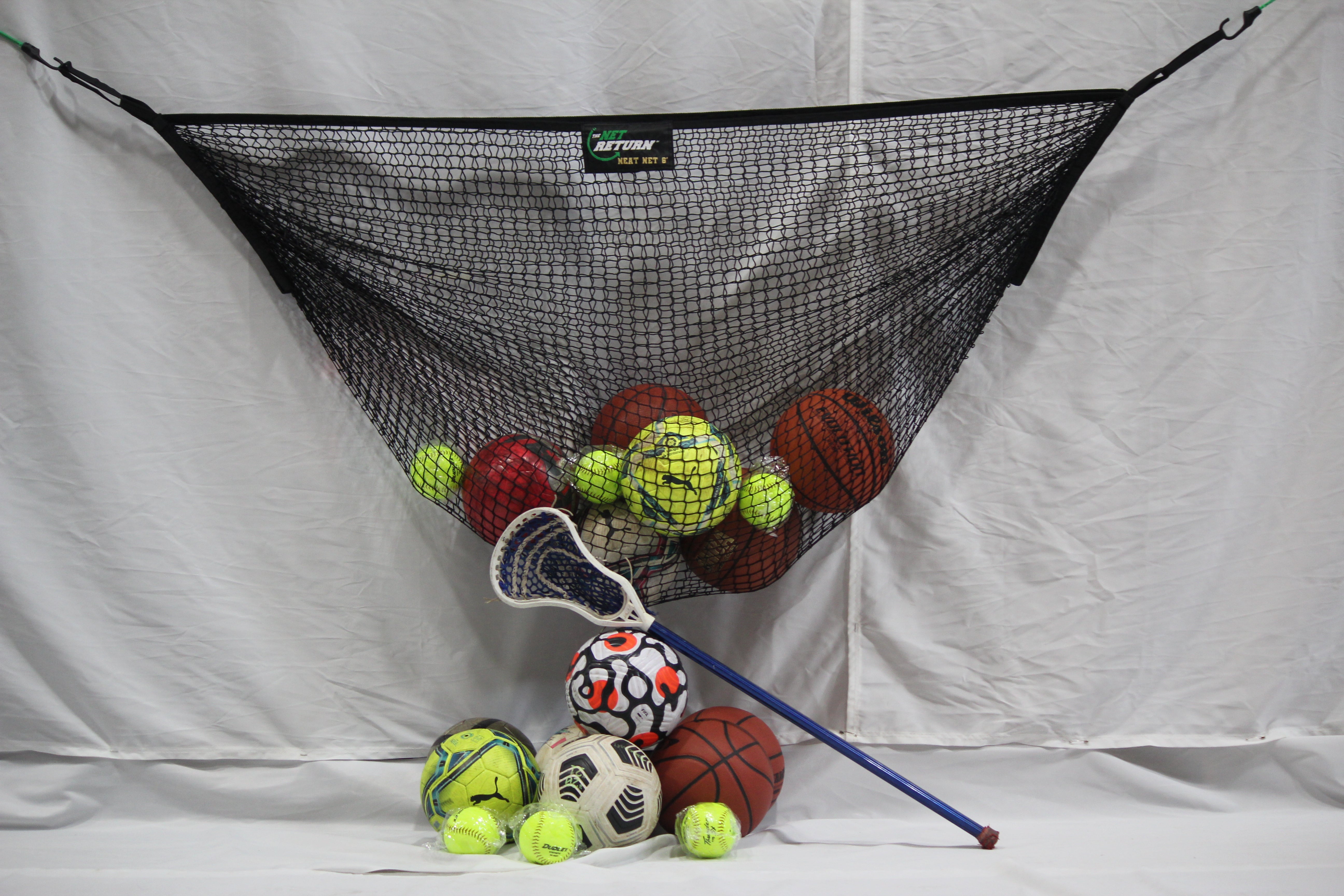 A variety of sports balls and a lacrosse stick stored in and around a net hanging against a white background.
