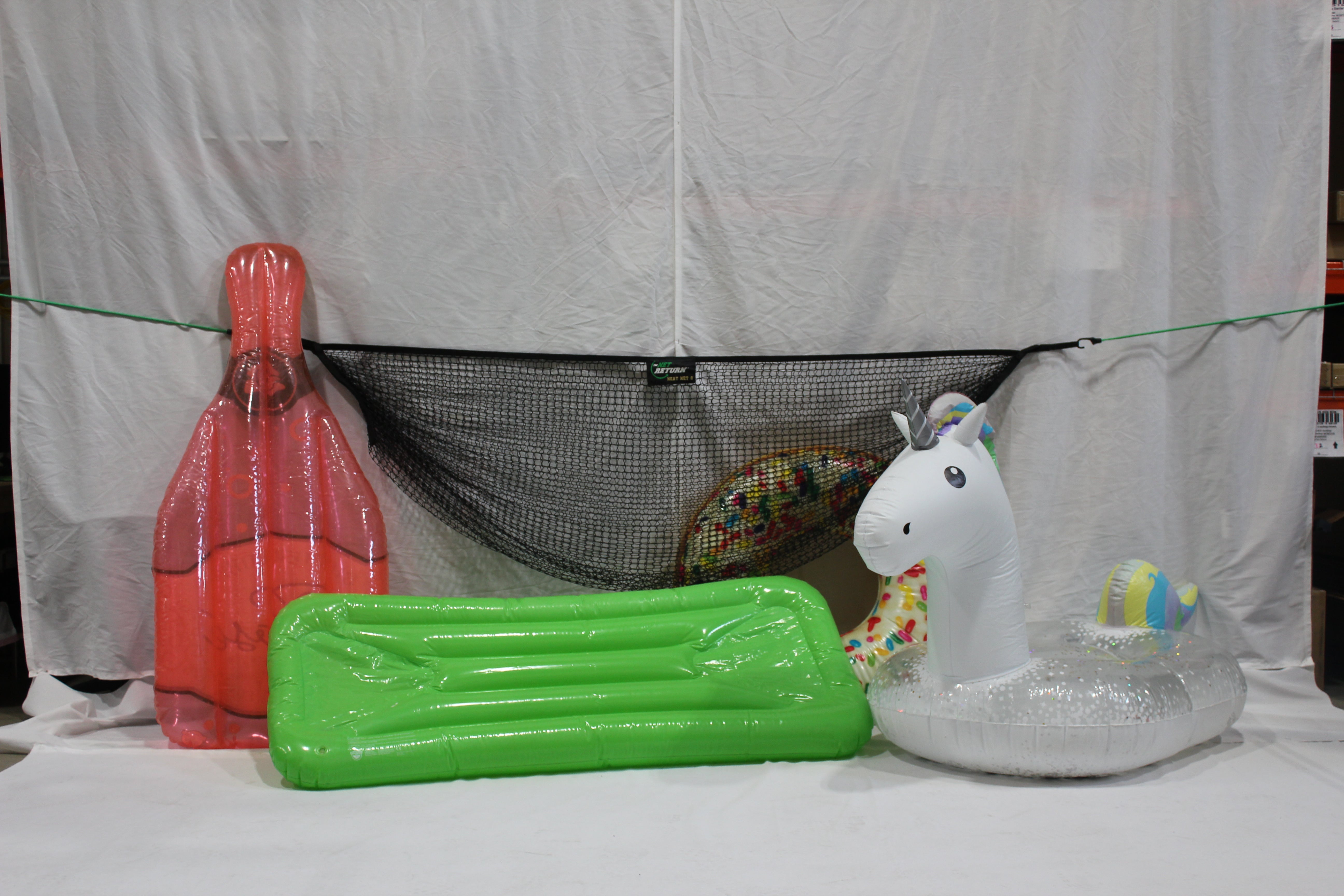 Three colorful inflatable pool floats: a red swim fin, a green lounge, and a white unicorn, arranged in front of a white curtain with a black net.