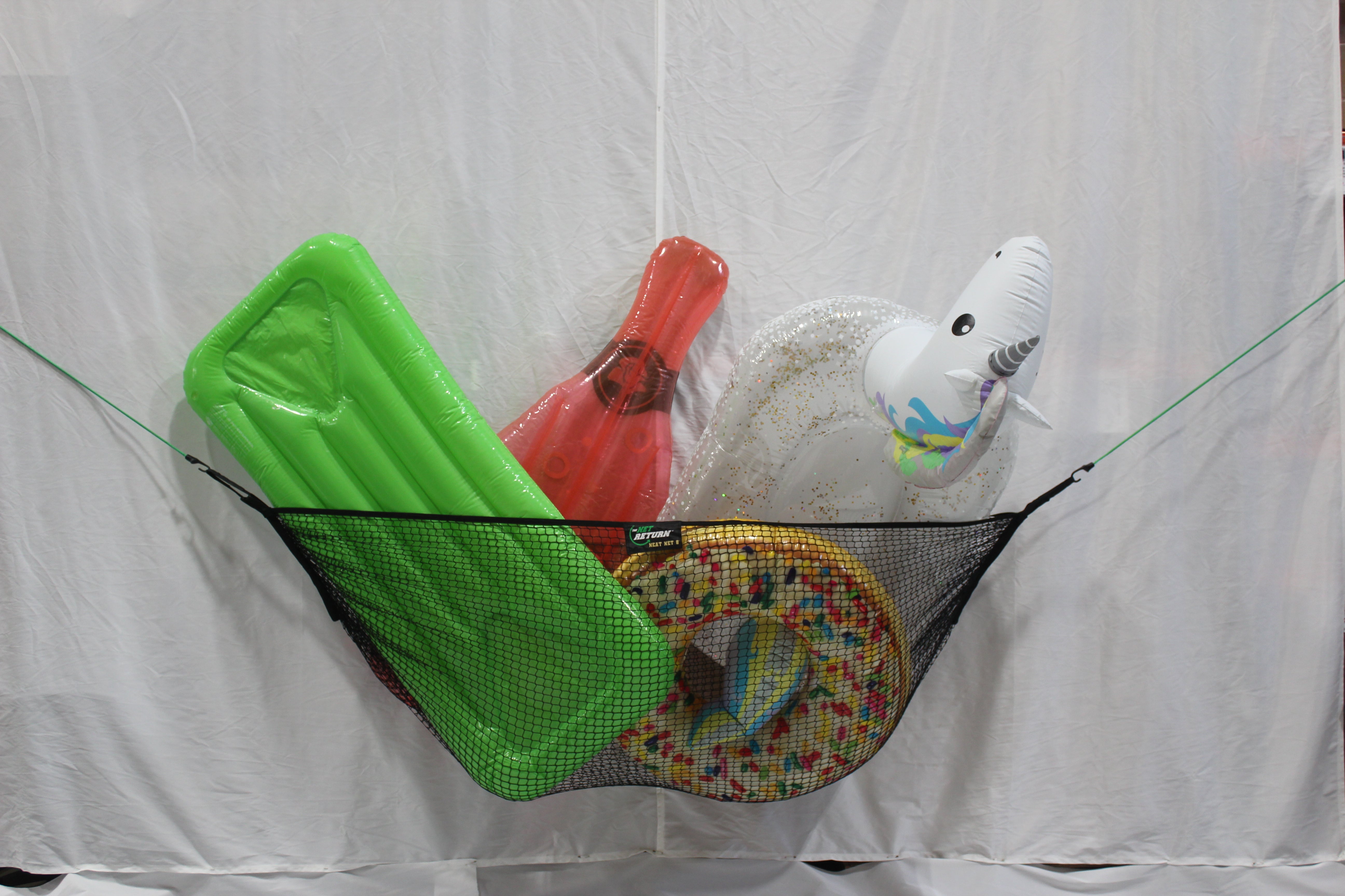 Inflatables including a green mattress, a red guitar, and a white unicorn with glitter, stored in a black mesh hammock against a white backdrop.
