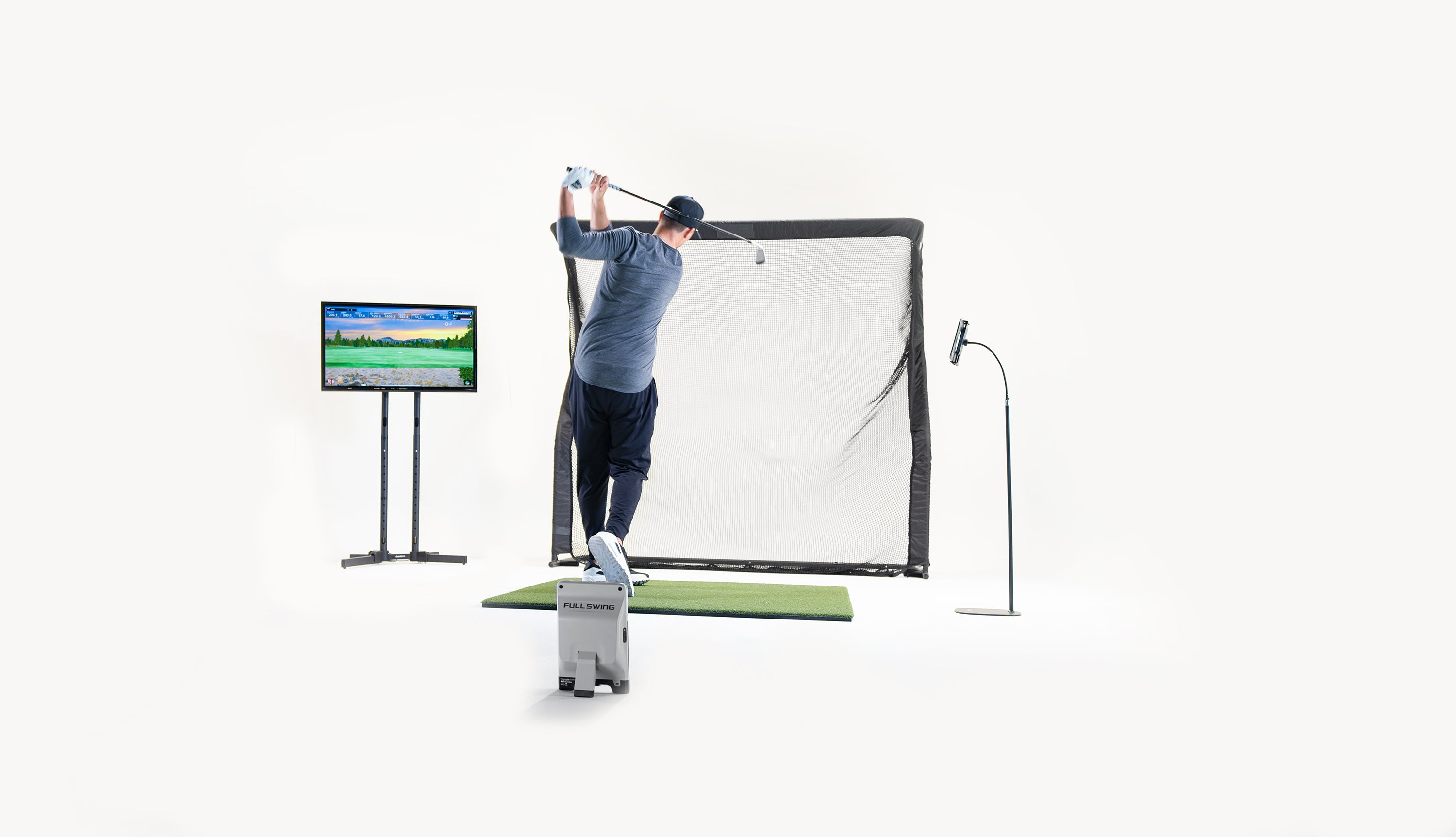 Person practicing golf swing into a net setup indoors, with a monitor displaying a golf course image nearby.