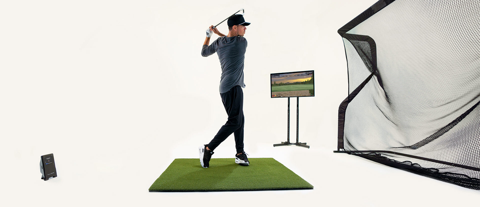 Person swinging a golf club in an indoor setup with a golf mat, net, and monitor displaying a landscape.