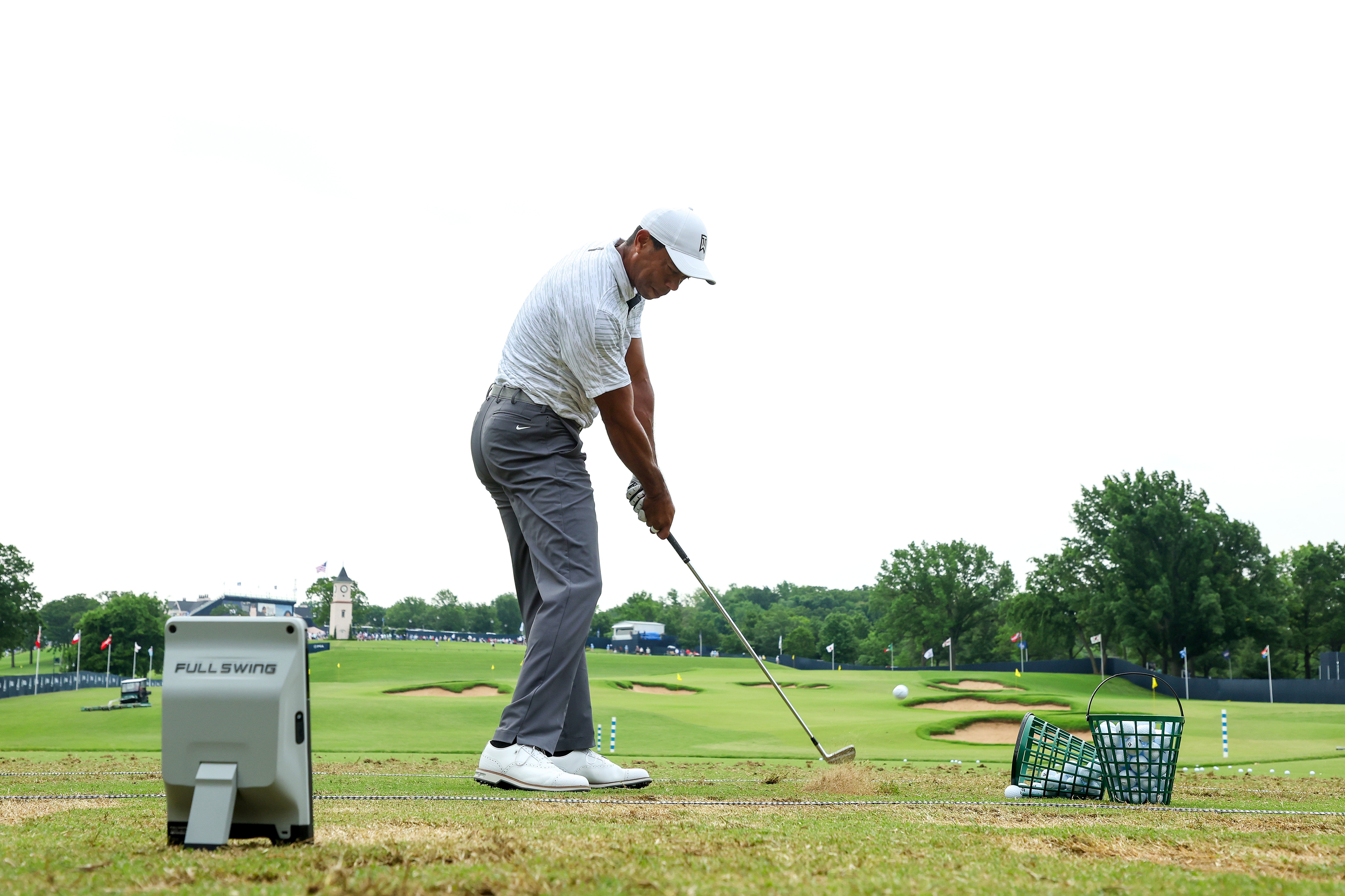 A golfer practices swinging at a driving range, with a basket of golf balls nearby and a monitoring device on the ground. Trees and a building are visible in the background.