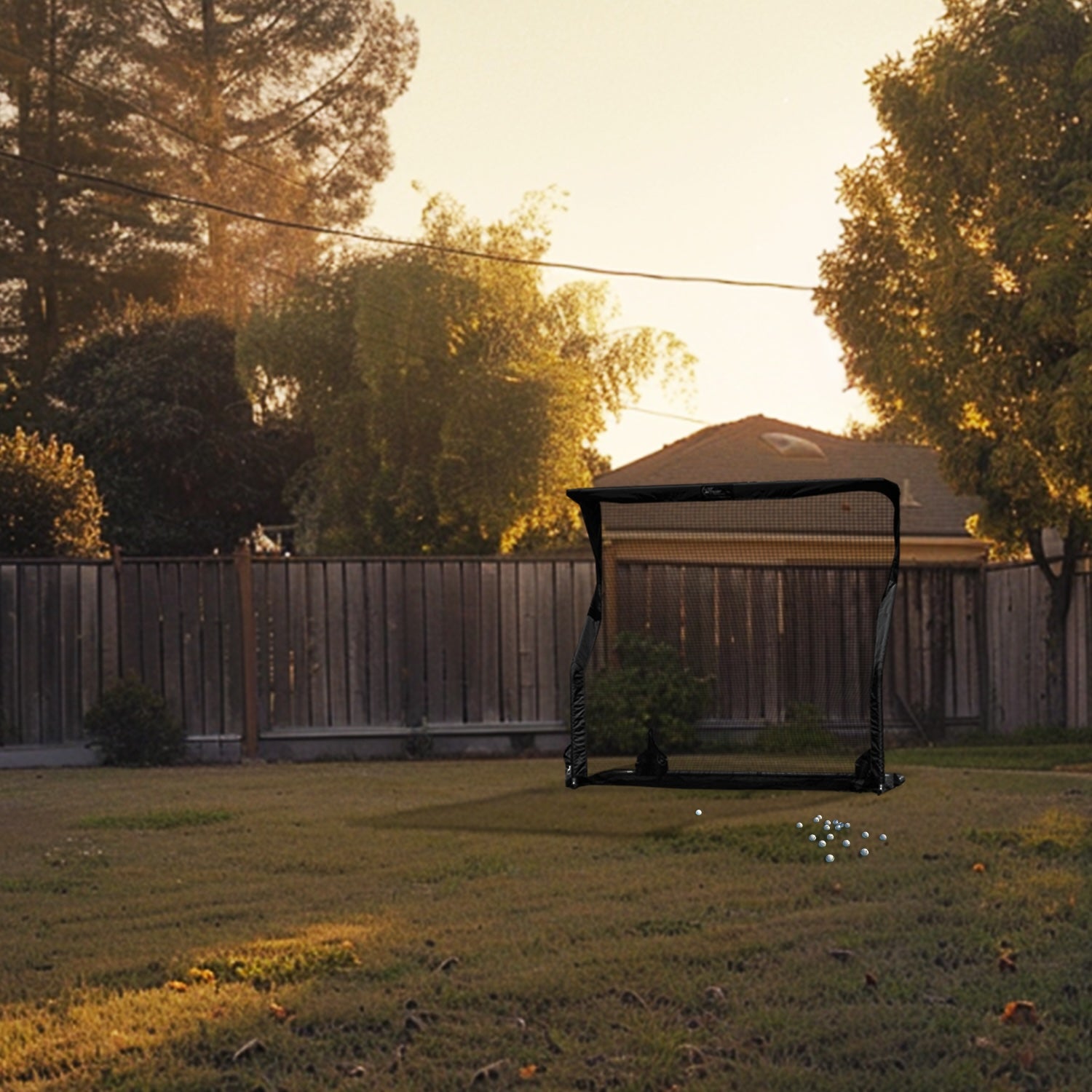 Golf net set up in a backyard during sunset, with scattered golf balls on the grass. A wooden fence and trees in the background.