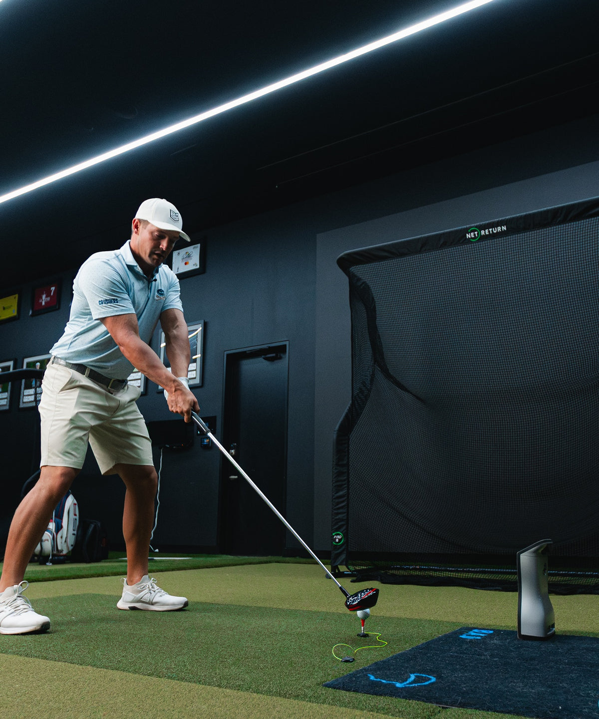 A golfer prepares to swing in an indoor practice area with a net and a monitor.