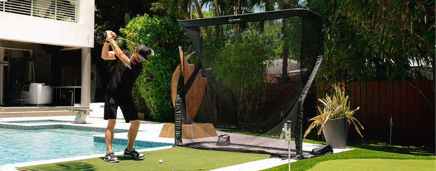 A person in a black outfit practices golf swings on a green mat near a pool, using a net for capturing golf balls.