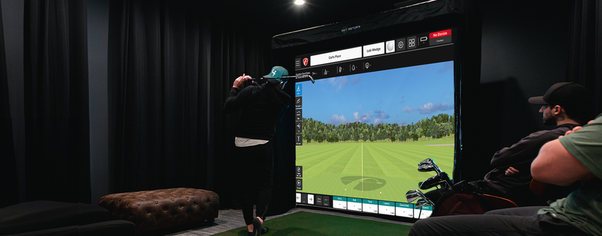 Person using an indoor golf simulator, swinging a club while another person watches. The screen displays a golf course.