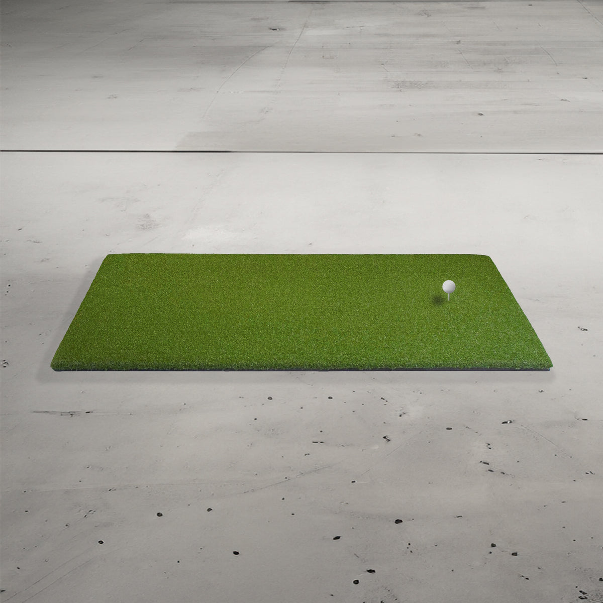 A green artificial turf mat on a concrete floor with a golf ball on a tee.