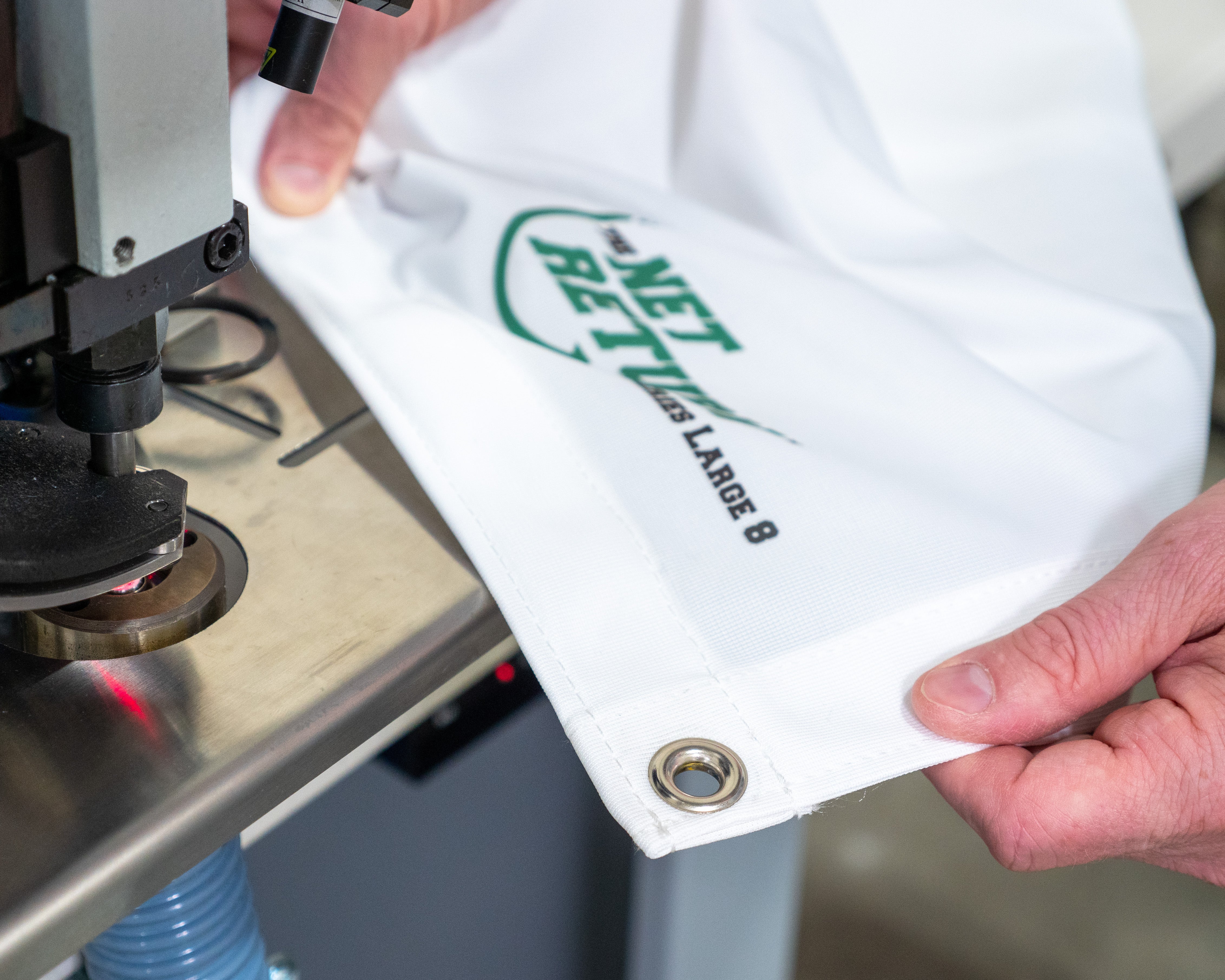 Close-up of a person sewing a white cloth with metal grommets, labeled "NETBET L LARG 8," using a sewing machine in a workshop.