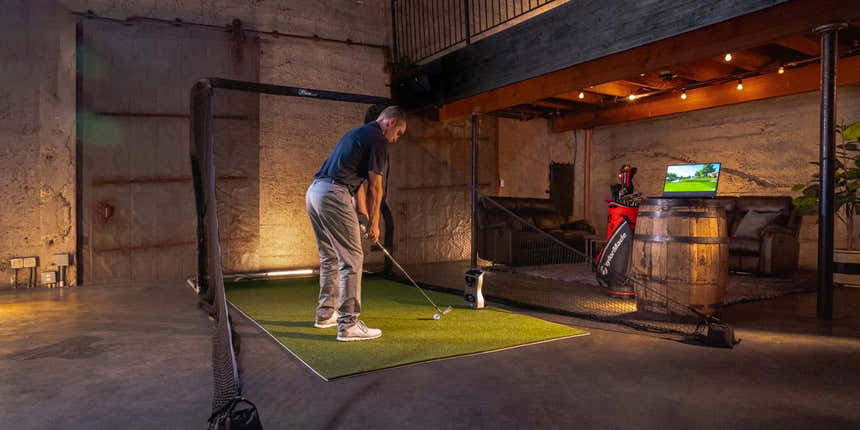 A person stands on an indoor golf simulator mat, preparing to swing. A laptop displays a golf course screen. The room has rustic decor with wooden beams and string lights.