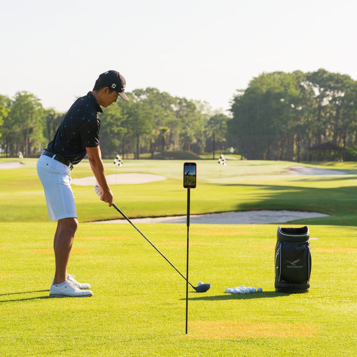 At the driving range, a golfer uses the GPOD by GPOD GOLF to mount his phone for easy golf swing video capture, while his golf bag sits nearby.