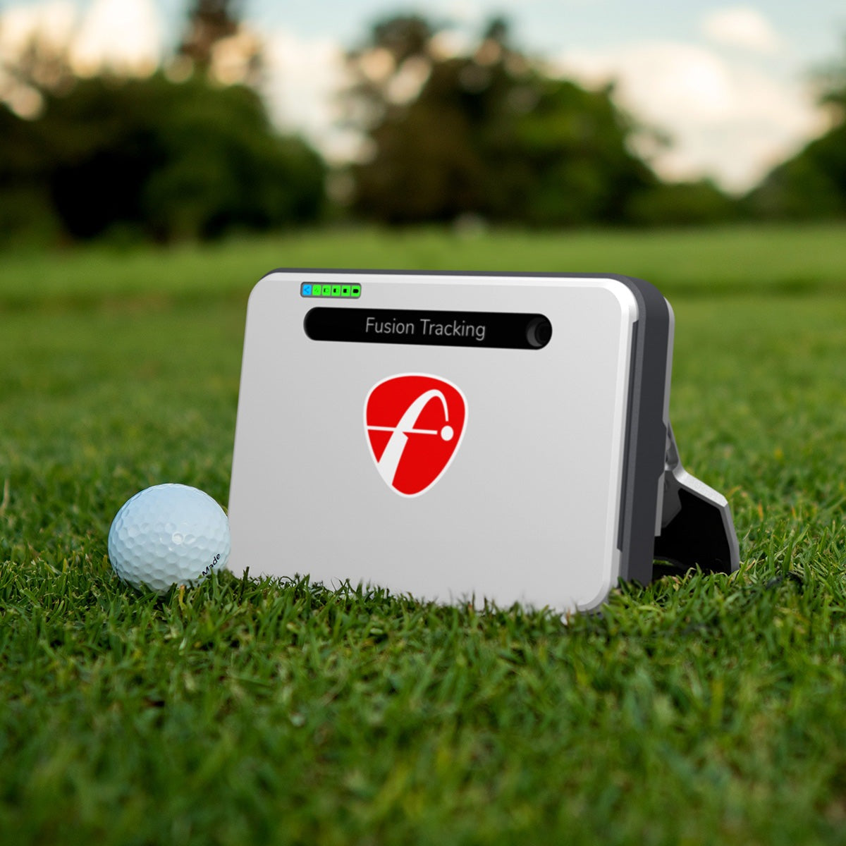 A white rectangular FlightScope Mevo+ device with a red logo and "Fusion Tracking" text rests on a grassy field beside a golf ball, capturing the essence of Simulated Golf Courses against a blurred backdrop of trees and sky.