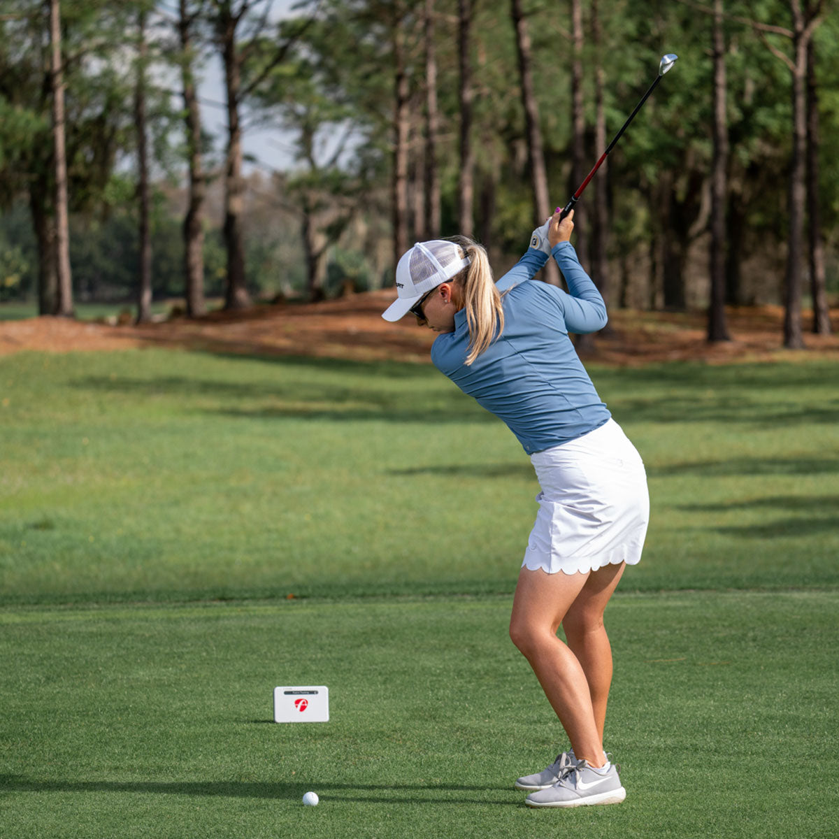 A person in a blue long-sleeve shirt, white skirt, and cap is swinging a golf club on a grassy course using FlightScope Mevo+ technology by FlightScope Golf. Trees line the background.