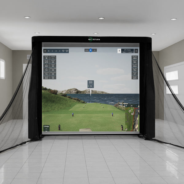 Indoor golf simulator with a large screen displaying a coastal golf course scene, surrounded by a net, in a spacious room with tiled flooring.