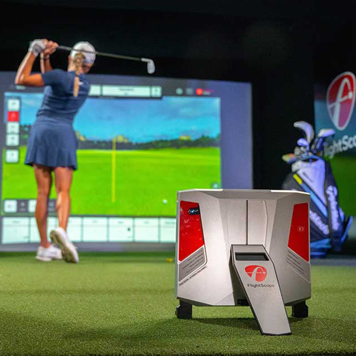 A person swings a golf club indoors with a simulation screen displaying immersive courses. In the foreground, a FlightScope X3 from FlightScope Golf uses Fusion Tracking technology to capture every swing detail.