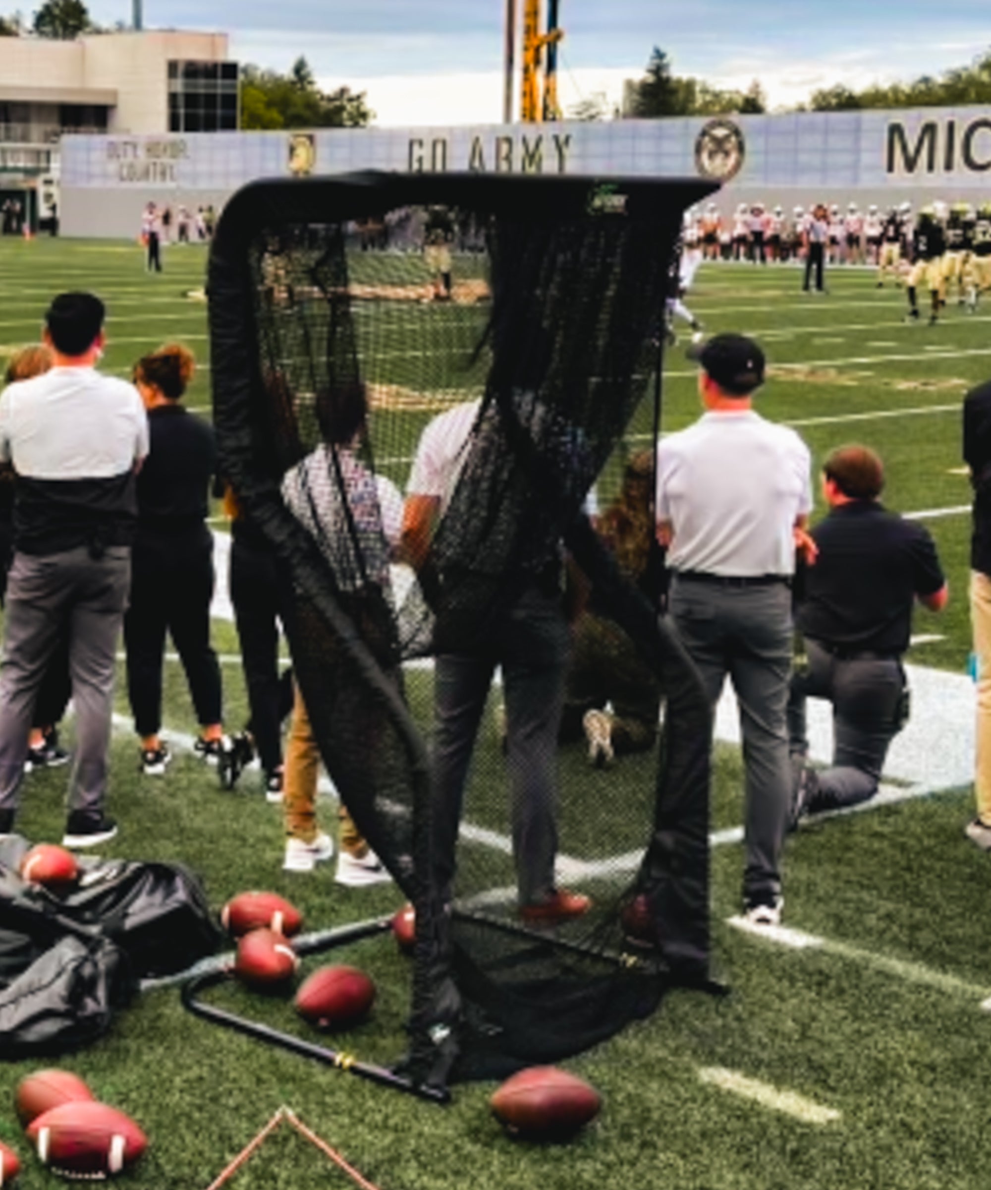 Football practice scene with people standing near a black net barrier on the field. Several footballs are on the ground. Stadium in the background.